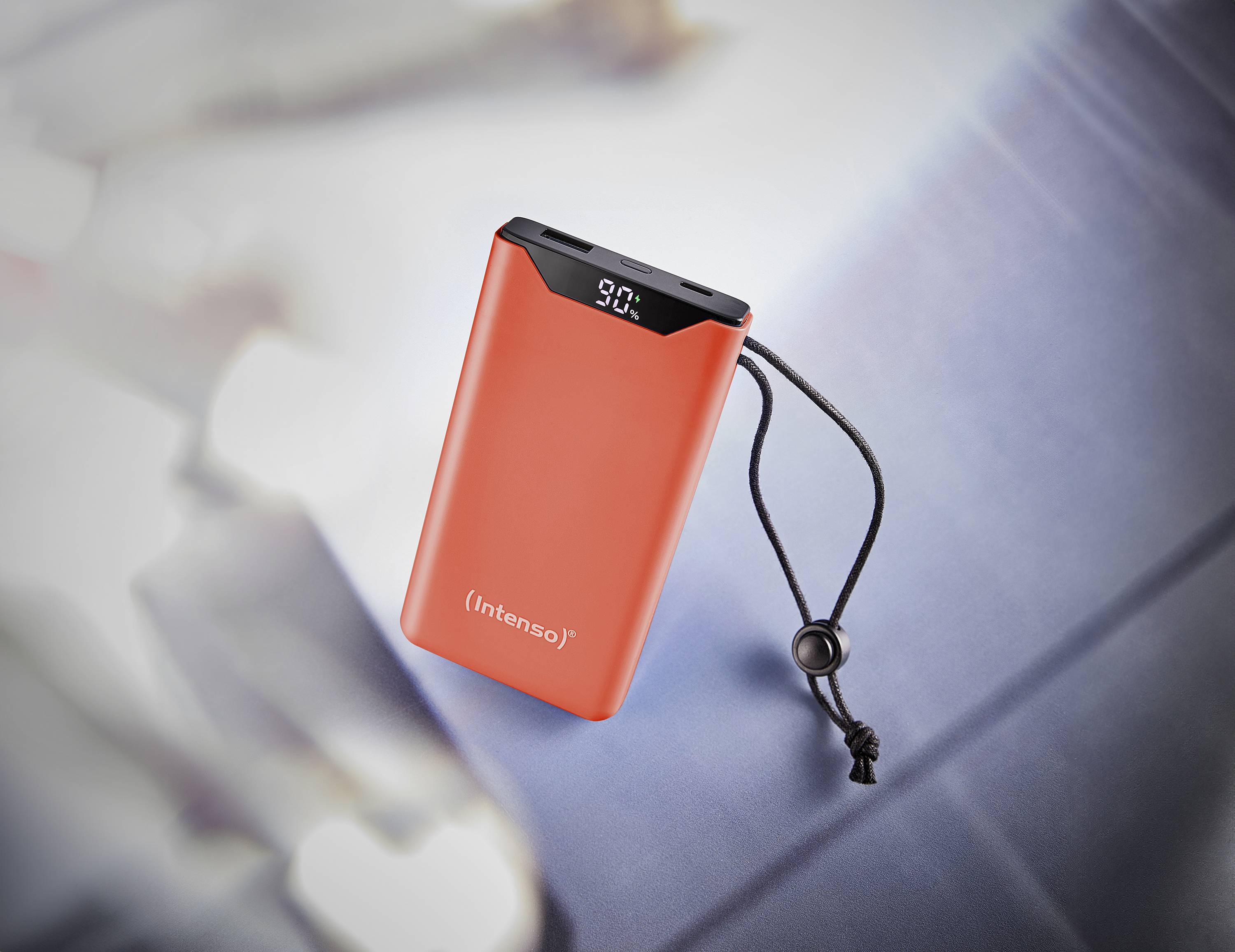 Rote Powerbank mit digitaler Anzeige '90%' und schwarzer Trageschlaufe, platziert auf einem metallischen Hintergrund.
