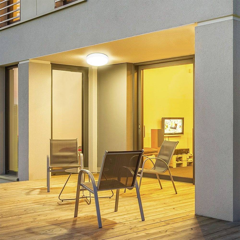 Ein moderner Balkon mit Holzdeck, zwei leeren Stühlen und einer kleinen Glastisch. Im Hintergrund beleuchtetes Zimmer eines Hauses.