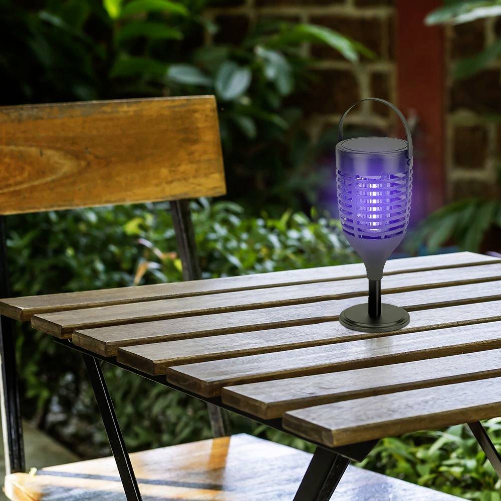 Ein elektrischer Insektenvernichter steht auf einem Holztisch im Freien, umgeben von Pflanzen und einem Holzzaun im Hintergrund.