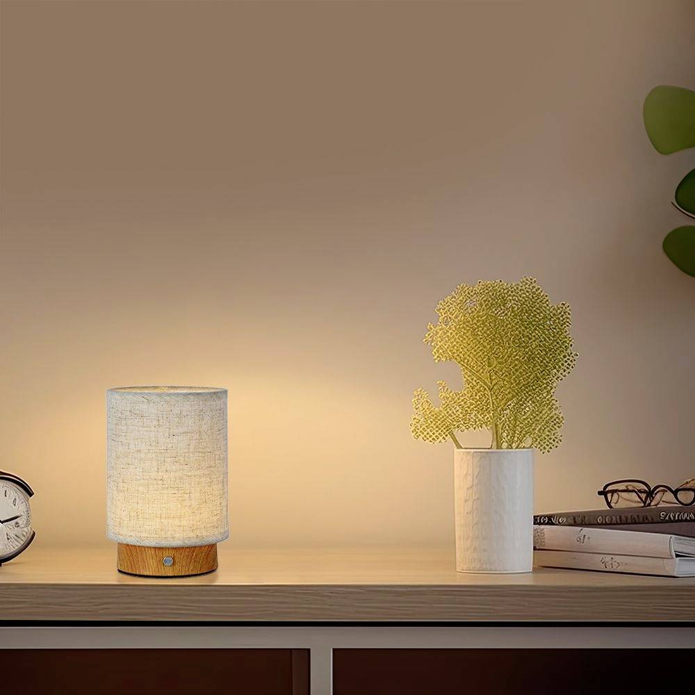 Eine Tischlampe mit hellem Schirm steht auf einem Holzregal neben einer Vase mit gelber Blume, im Hintergrund sind Bücher sichtbar.