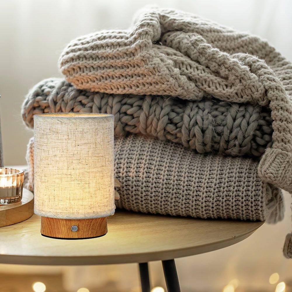 Tischlampe mit Holzfuß leuchtet warm, daneben gefaltete Strickdecken auf hellem Holztisch, gemütliche Atmosphäre.