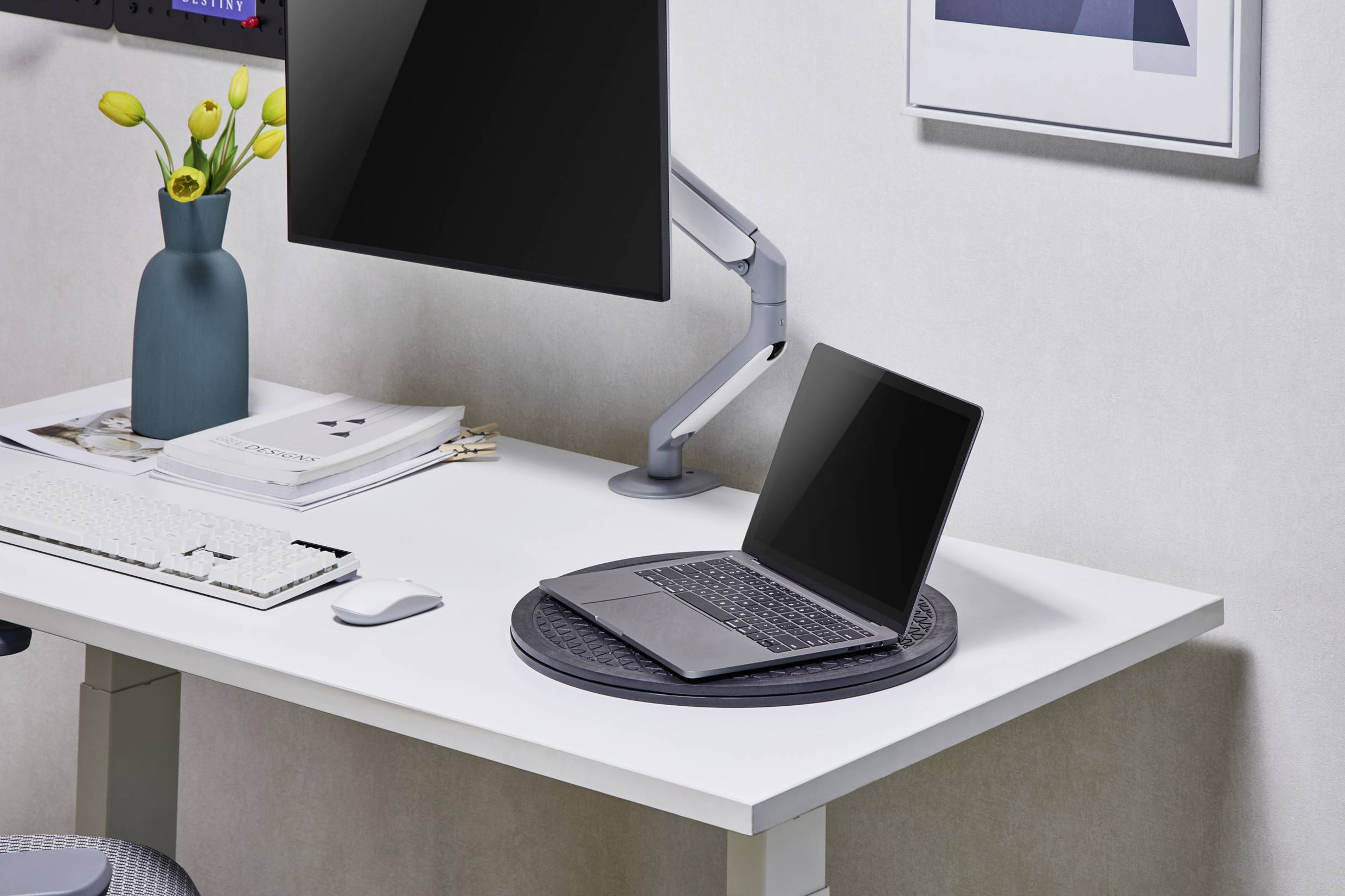 Ein moderner Arbeitsplatz mit einem Laptop auf einem Tisch, einem Monitor auf einem Standfuß, einer Tastatur und einer Vase mit gelben Blumen.