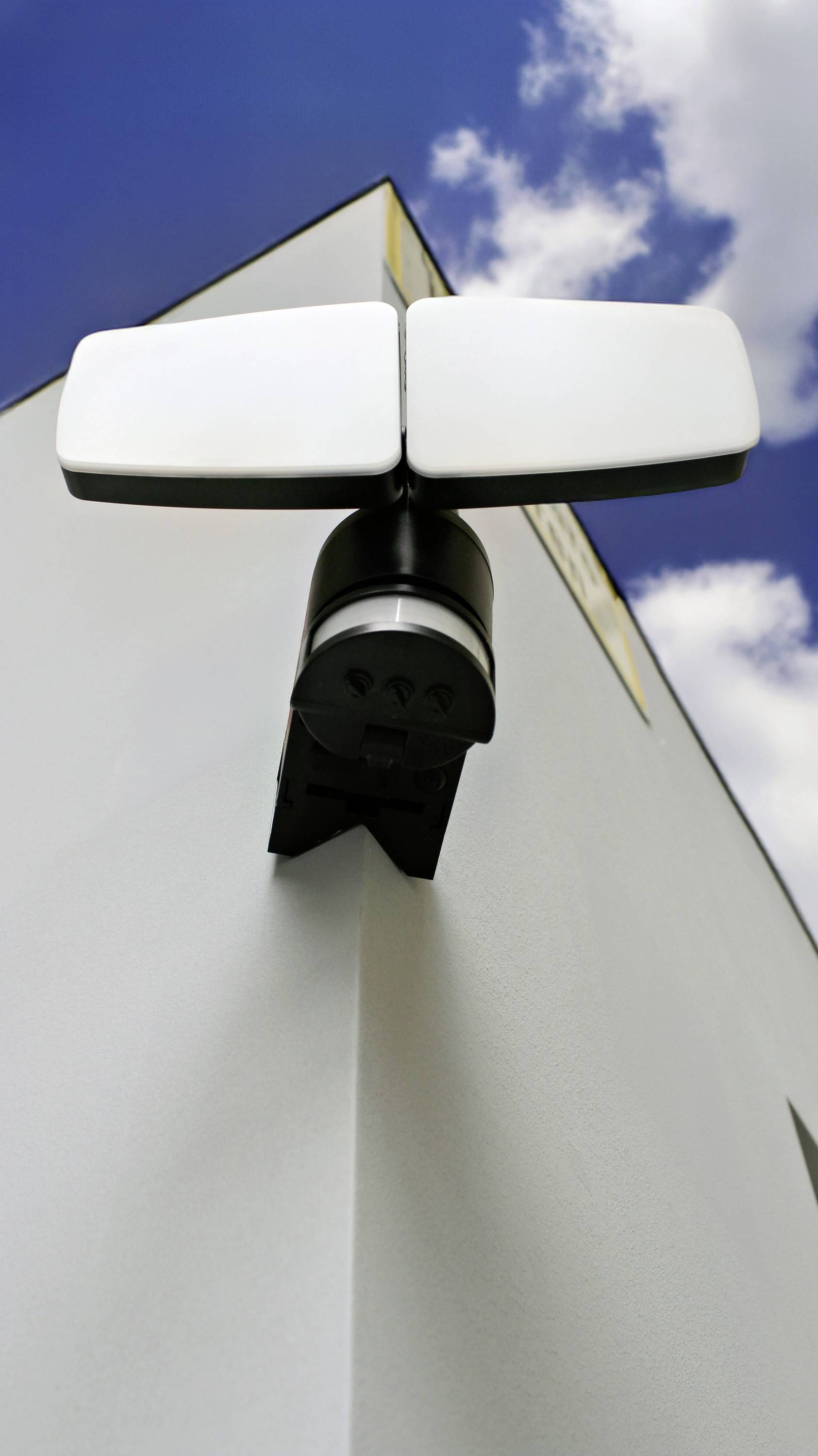 Eine Außenleuchte mit Bewegungsmelder ist an der Ecke eines Gebäudes angebracht, vor einem blauen Himmel mit einigen Wolken.