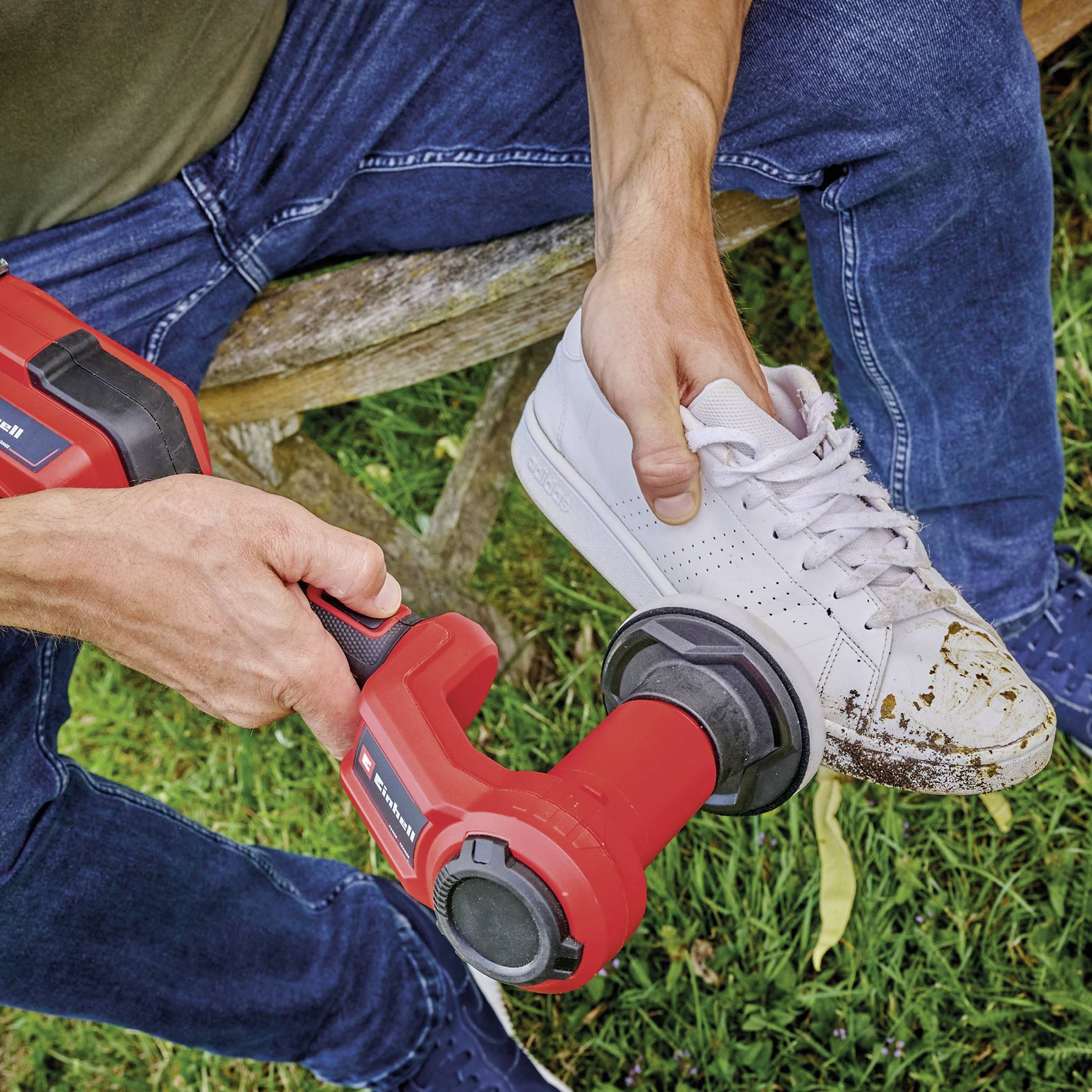 Eine Person reinigt einen weißen Sportschuh mit einem roten elektrischen Reinigungsgerät auf einer Wiese.
