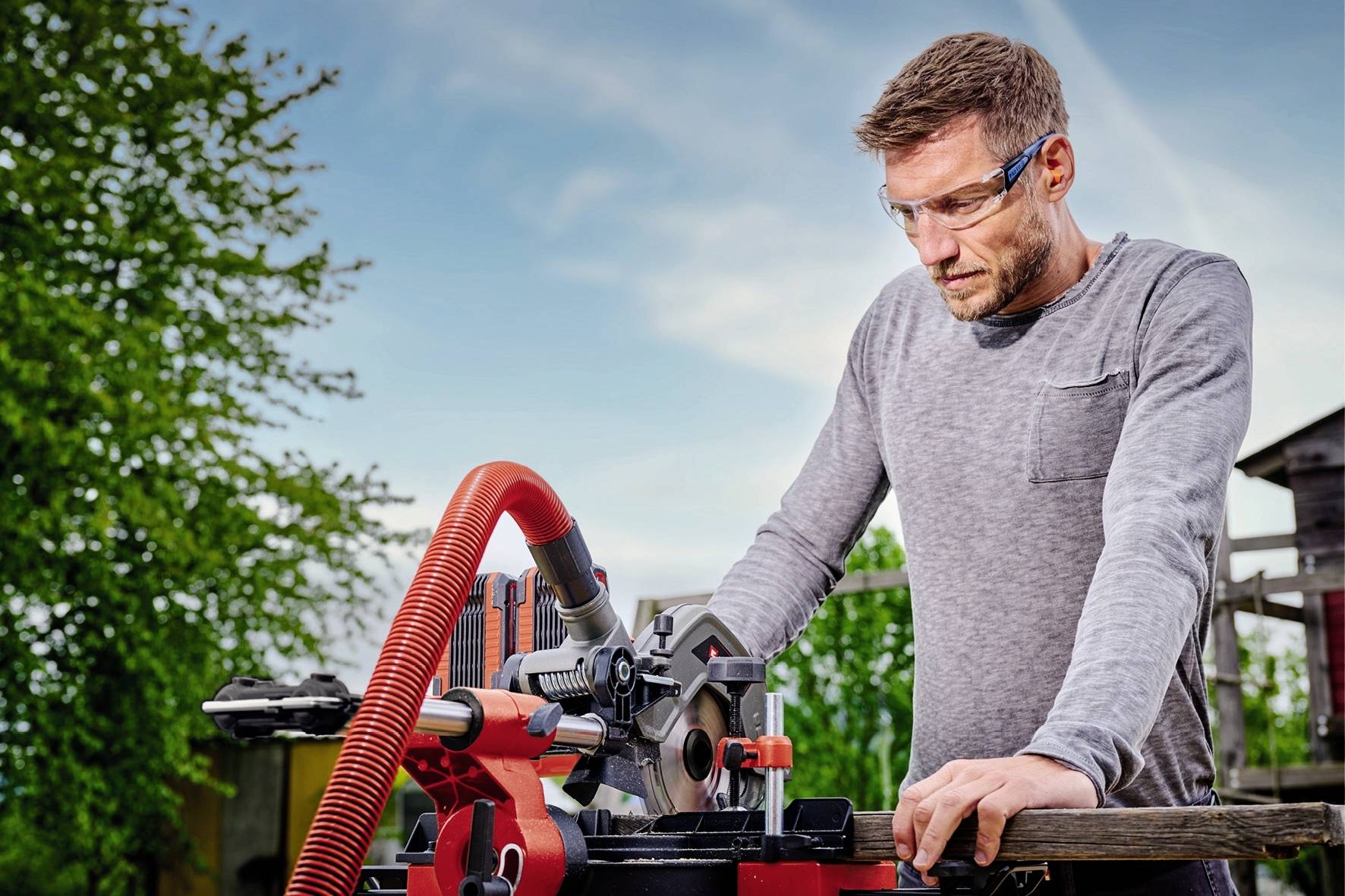 Ein Mann arbeitet an einer roten Kreissäge im Freien, trägt Schutzbrille und schaut konzentriert auf das Holz, das er schneidet.