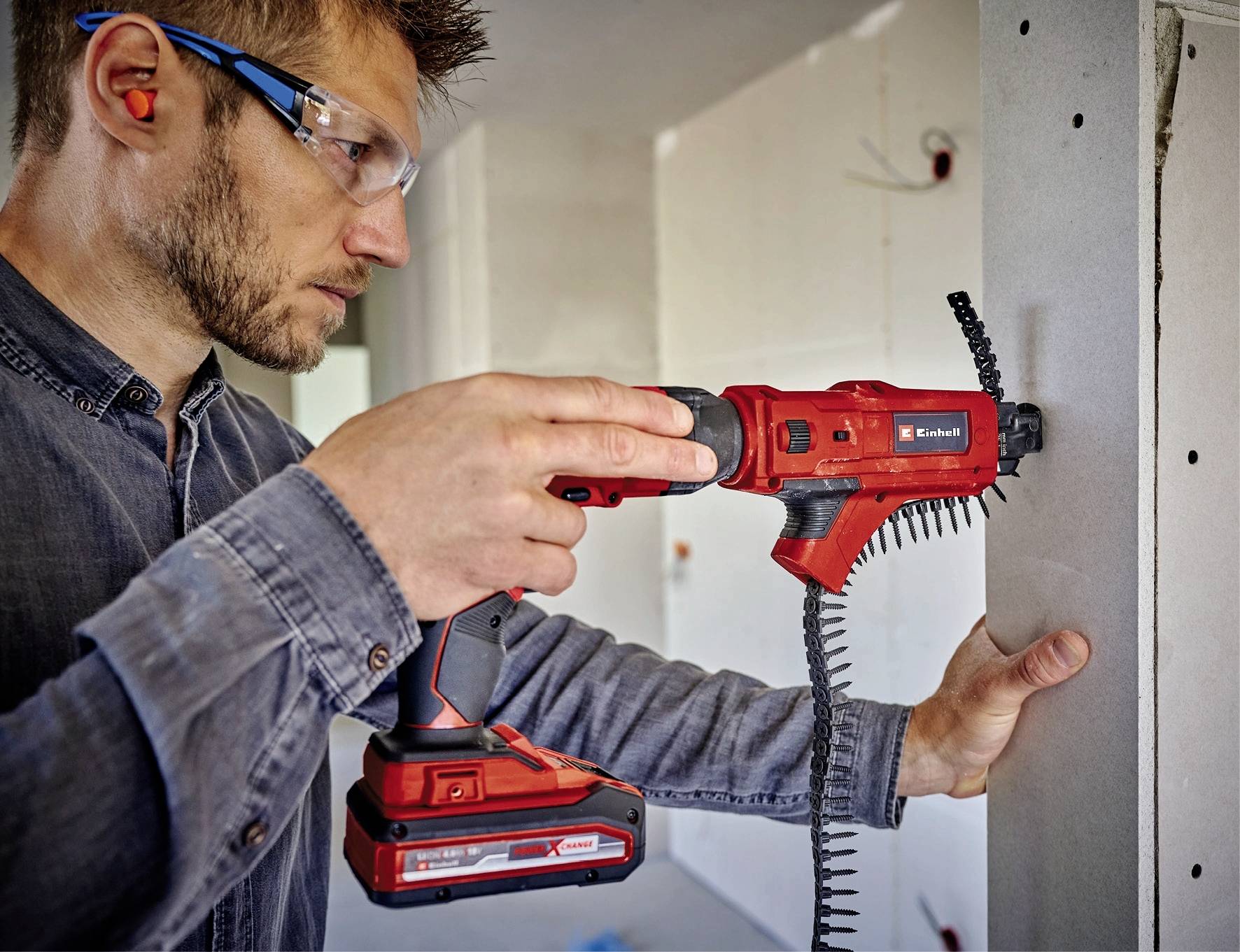 Ein Mann bohrt mit einer roten Bohrmaschine Löcher in eine Wand. Er trägt Schutzbrille und Ohrstöpsel. Im Hintergrund ist ein Raum zu sehen.