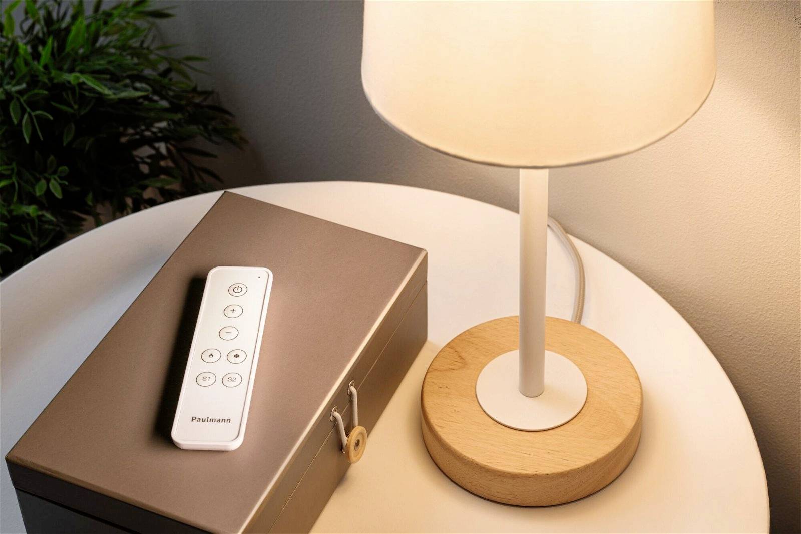 Weiße Fernbedienung auf einem braunen Kasten neben einer leuchtenden Tischlampe auf einem runden Tisch.