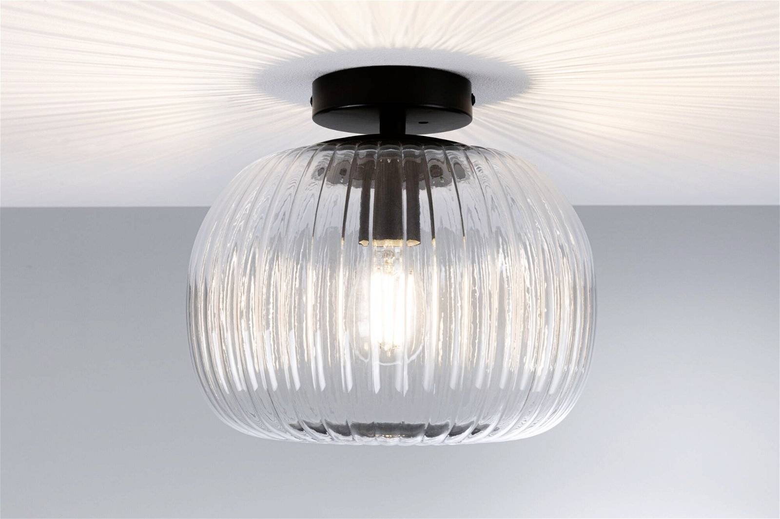 Deckenleuchte mit klarem geripptem Glas, das Licht gleichmäßig im Raum streut. Schwarzer Sockel betont das moderne Design.
