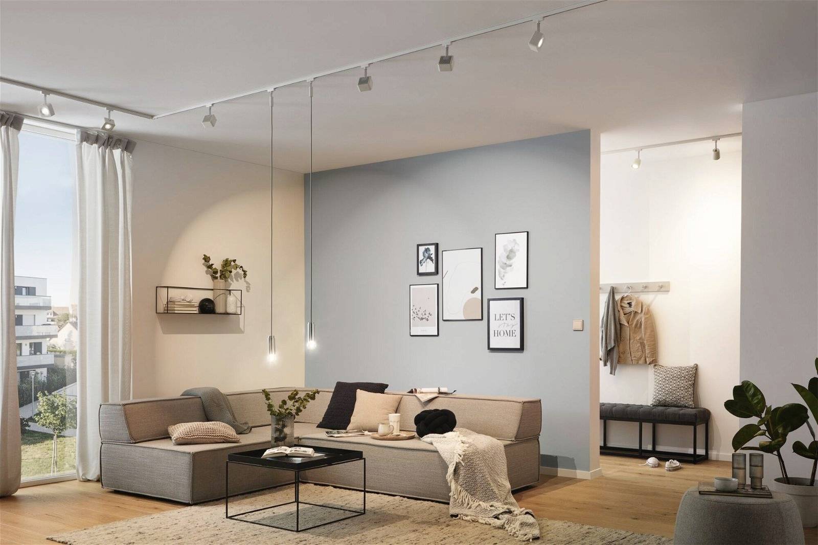 Helles Wohnzimmer mit Ecksofa, kleinem Couchtisch und Bilderwand. Fenster mit Tageslicht, Pflanzen und Kleidungsstücken im Hintergrund.