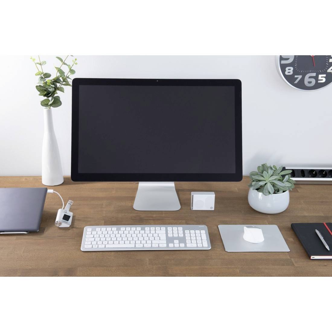 Ein aufgeräumter Schreibtisch mit einem großen Monitor, Tastatur, Maus und Pflanzen. Uhr an der Wand zeigt kurz nach neun.