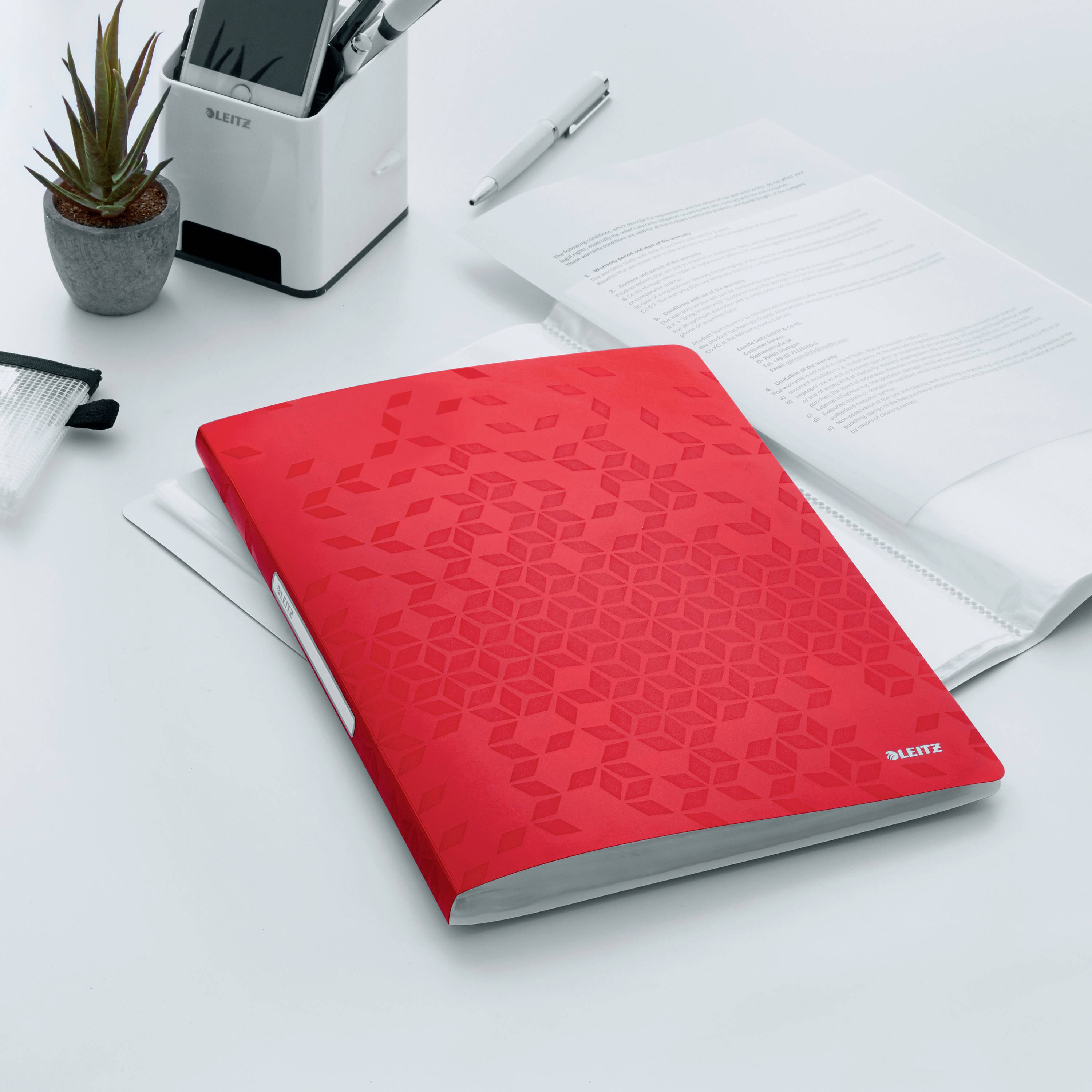 Ein roter Ordner liegt auf einem Schreibtisch neben einem Dokument, einem Stift und einem Stifthalter mit Pflanzen, in einem Büro.