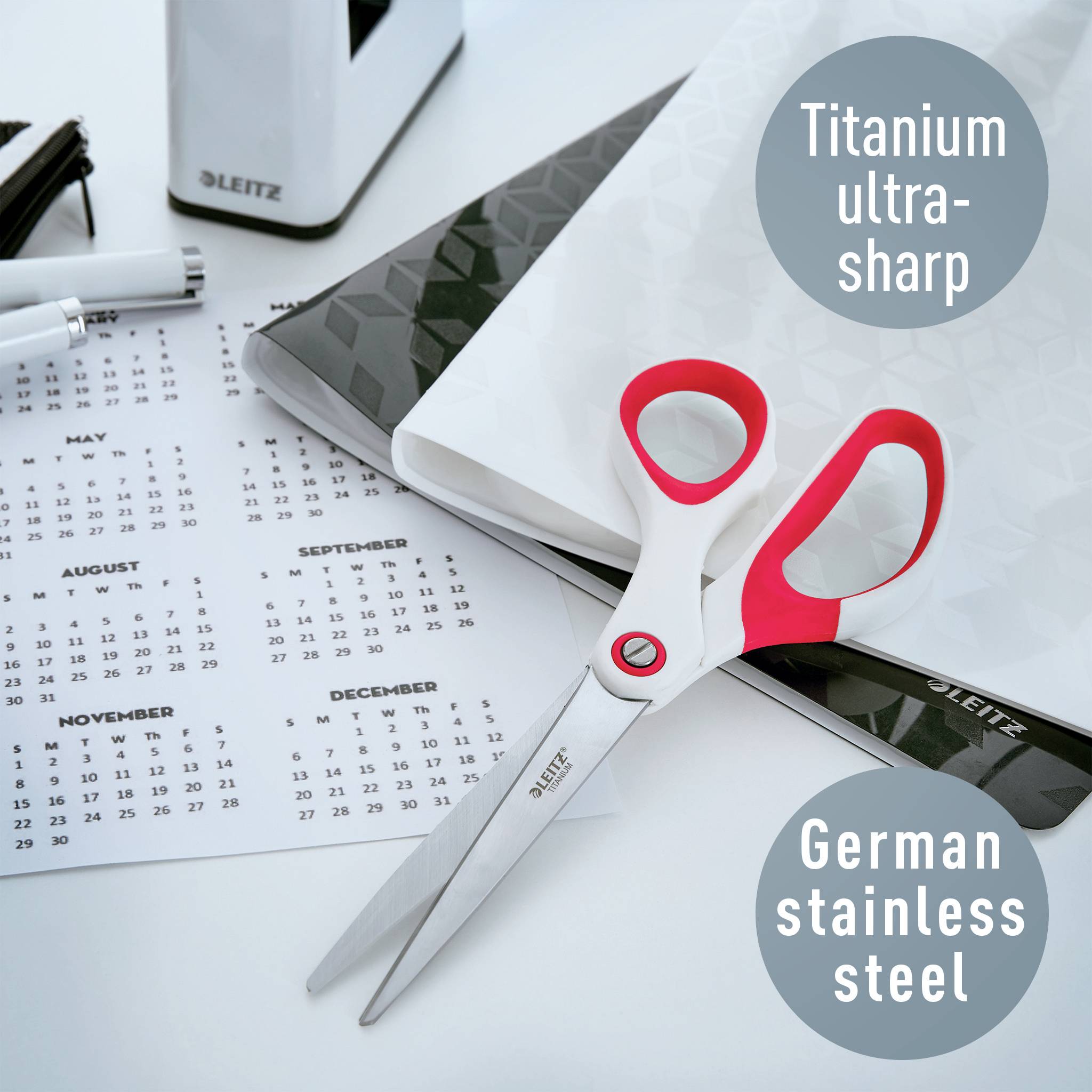 Beschreibt eine Schere mit rotem Griff auf einem Schreibtisch. Im Hintergrund sind ein Kalender und Bürogegenstände zu sehen. Aufschriften: 'Titanium ultra-sharp', 'German stainless steel'.