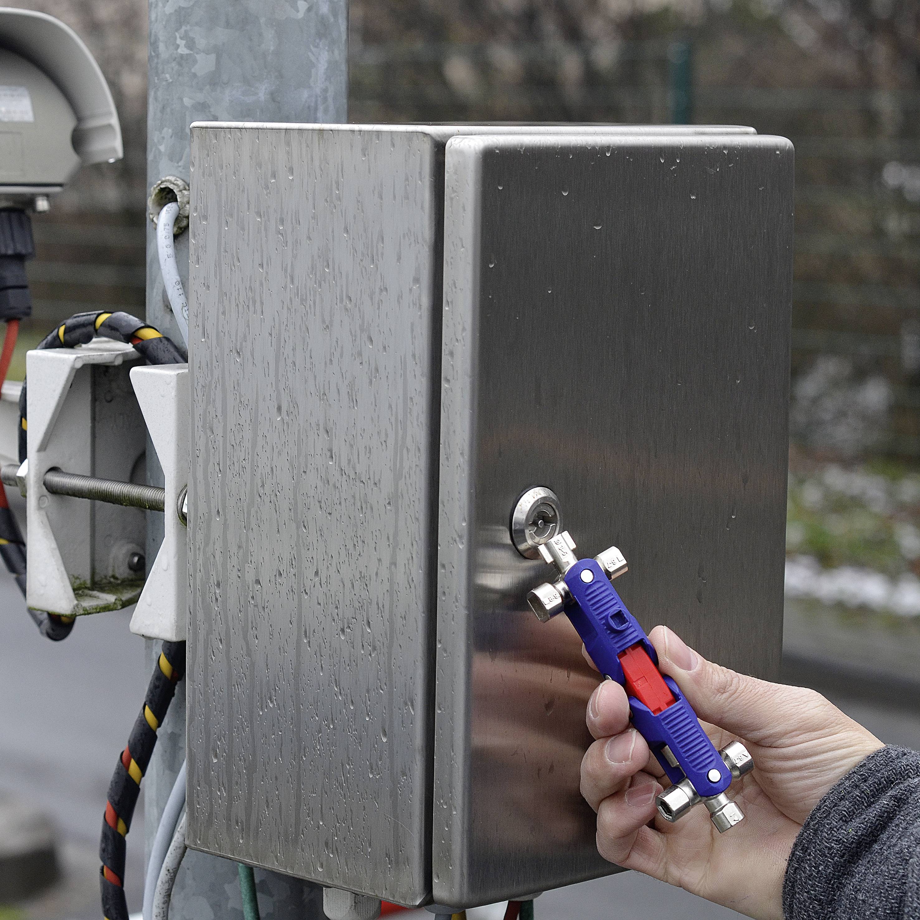 Hand hält Mehrfachschlüssel, öffnet silbernen Metallschaltkasten auf einem Pfeiler mit Kabeln. Draußen, trübes Wetter.