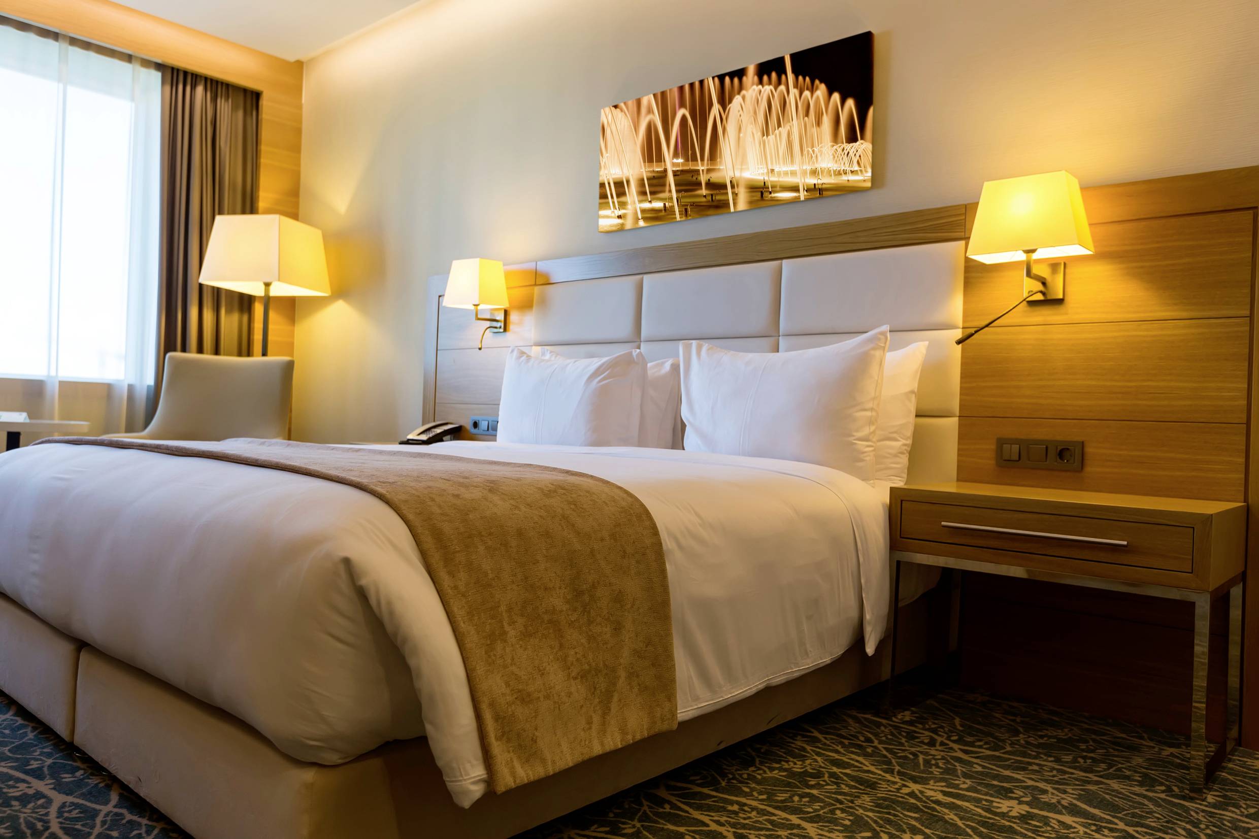 Geräumiges Hotelzimmer mit Doppelbett, zwei Nachttischen mit Lampen, einem Fenster mit Vorhang und einem modernen Kunstwerk an der Wand.