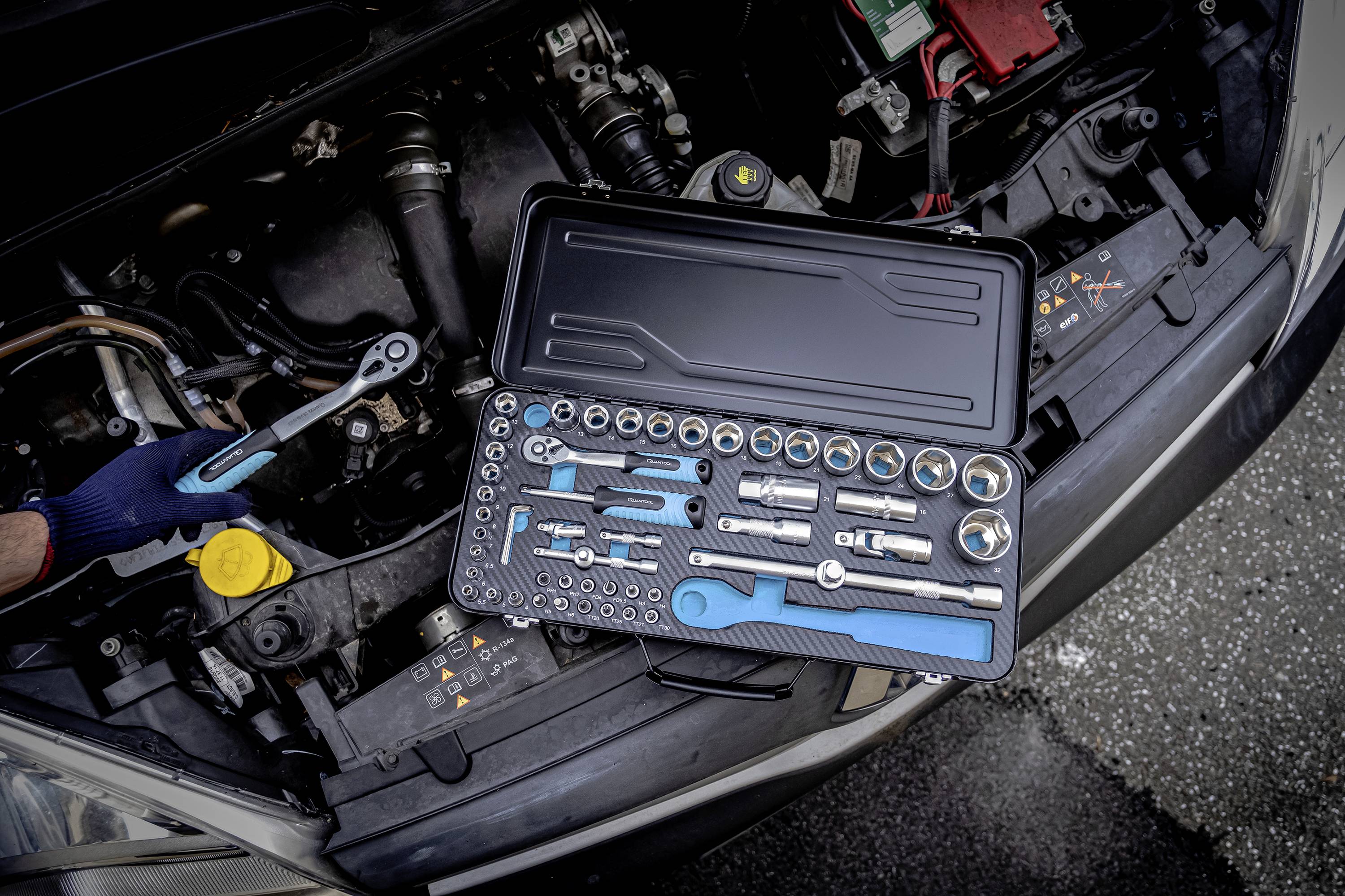 Kfz-Mechaniker benutzt Ratsche am geöffneten Motorraum eines Autos. Daneben liegt ein Steckschlüsselsatz mit verschiedenen Aufsätzen.