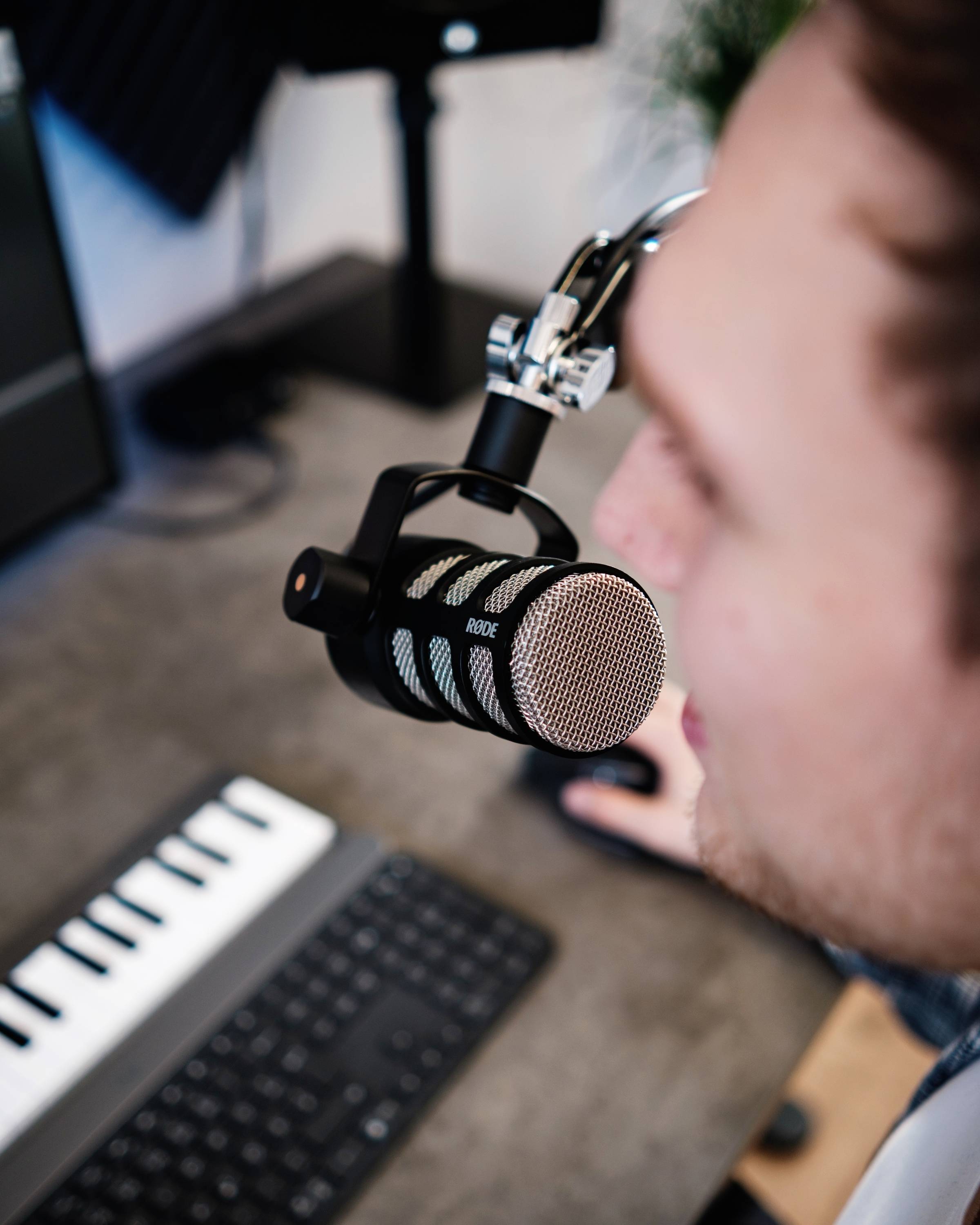 Ein Mensch spricht in ein Mikrofon in einem Studio. Im Vordergrund sind eine Computertastatur und ein MIDI-Keyboard zu sehen.