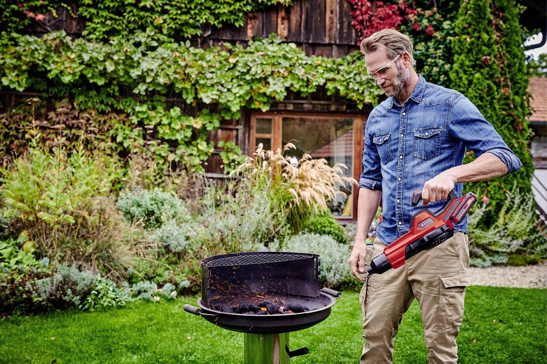 Ein Mann im Garten entzündet mit einem elektrischen Grillanzünder Holzkohle in einem runden Grill. Im Hintergrund sind viele Pflanzen.