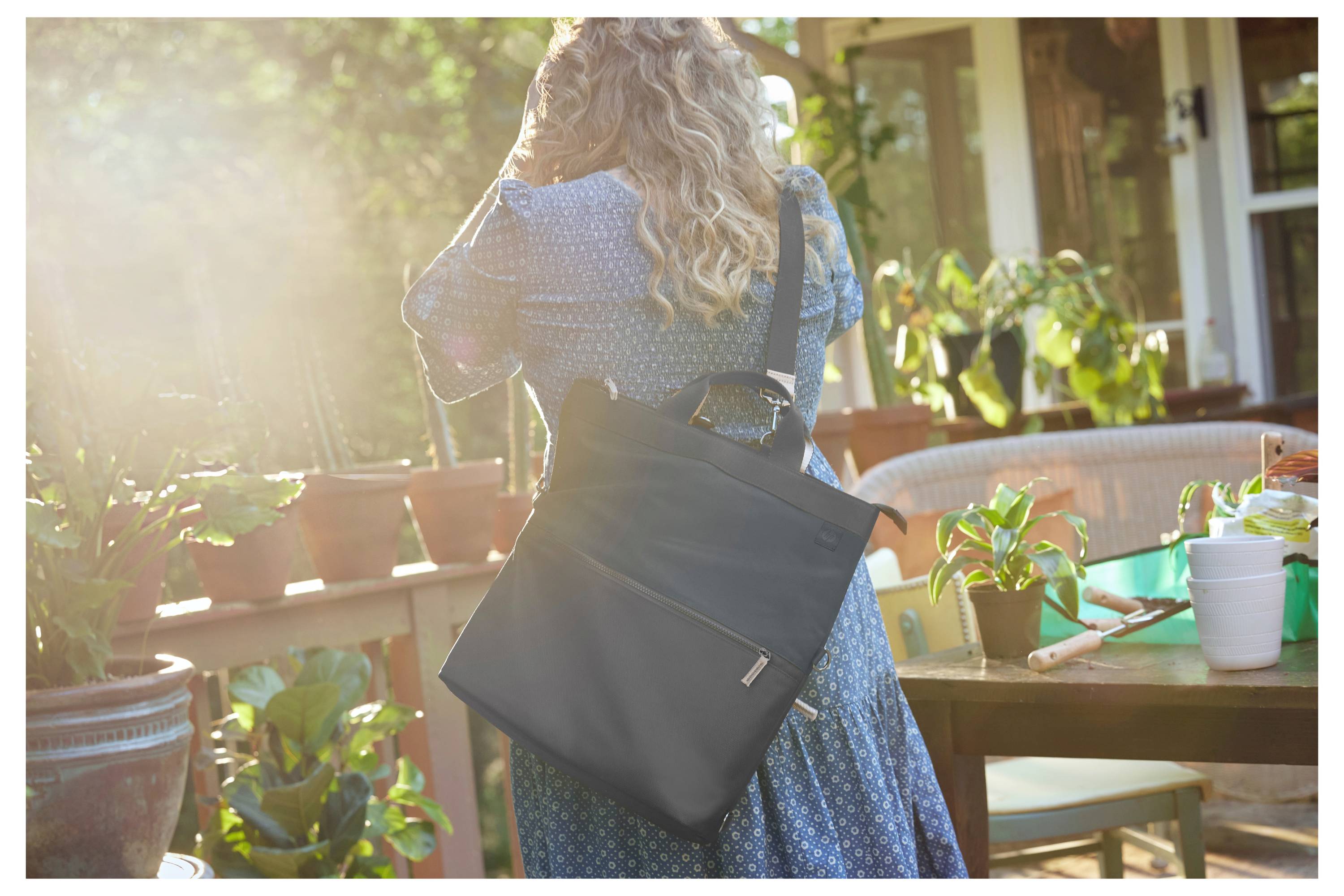 Frau in blauem Kleid mit schwarzer Tasche auf Terrasse mit Pflanzen bei Sonnenschein.