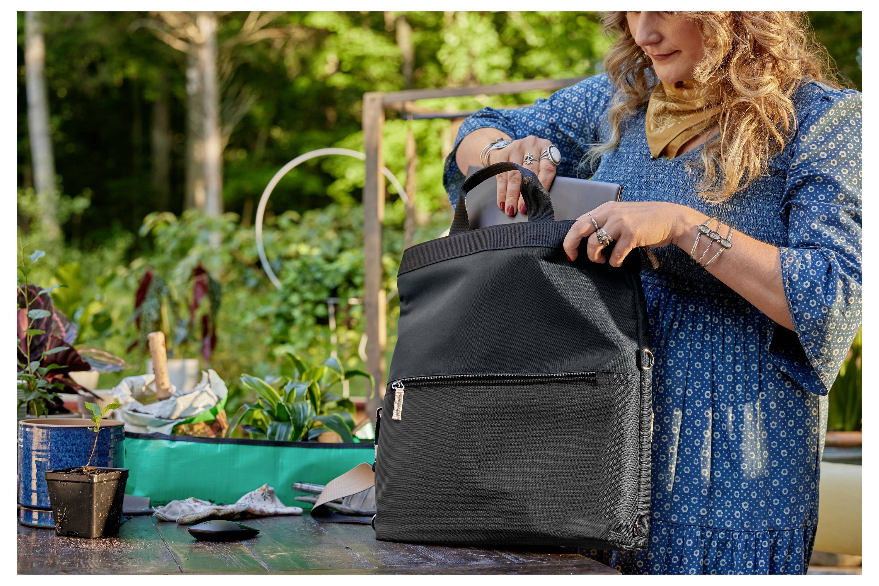 Eine Frau im blauen Kleid steht an einem Gartentisch und packt einen schwarzen Rucksack aus. Im Hintergrund sind Pflanzen sichtbar.