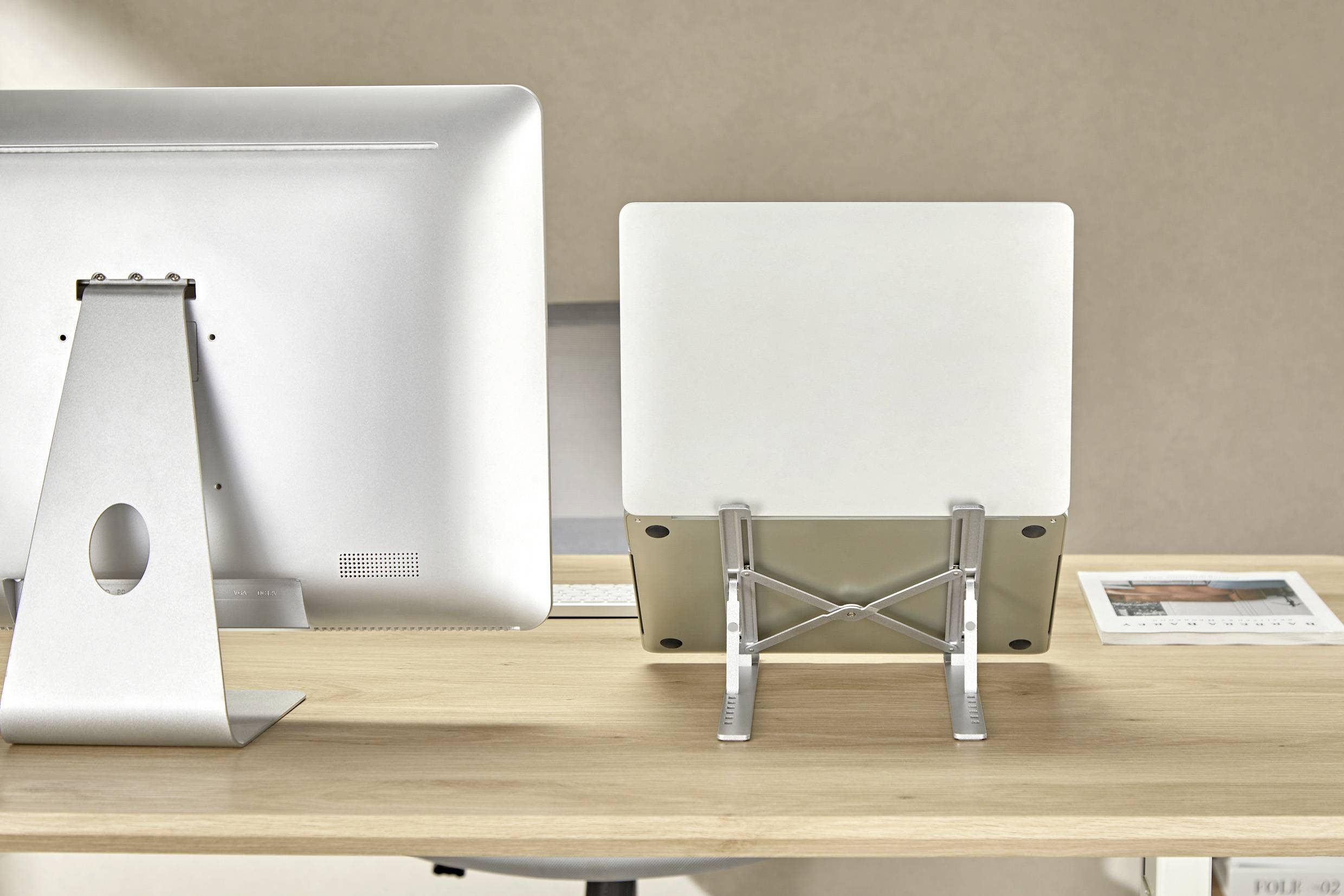 Ein minimalistischer Schreibtisch mit einem Computermonitor, einem Laptop auf einem Ständer und einem gerahmten Foto auf der rechten Seite, vor einem neutralen Hintergrund.
