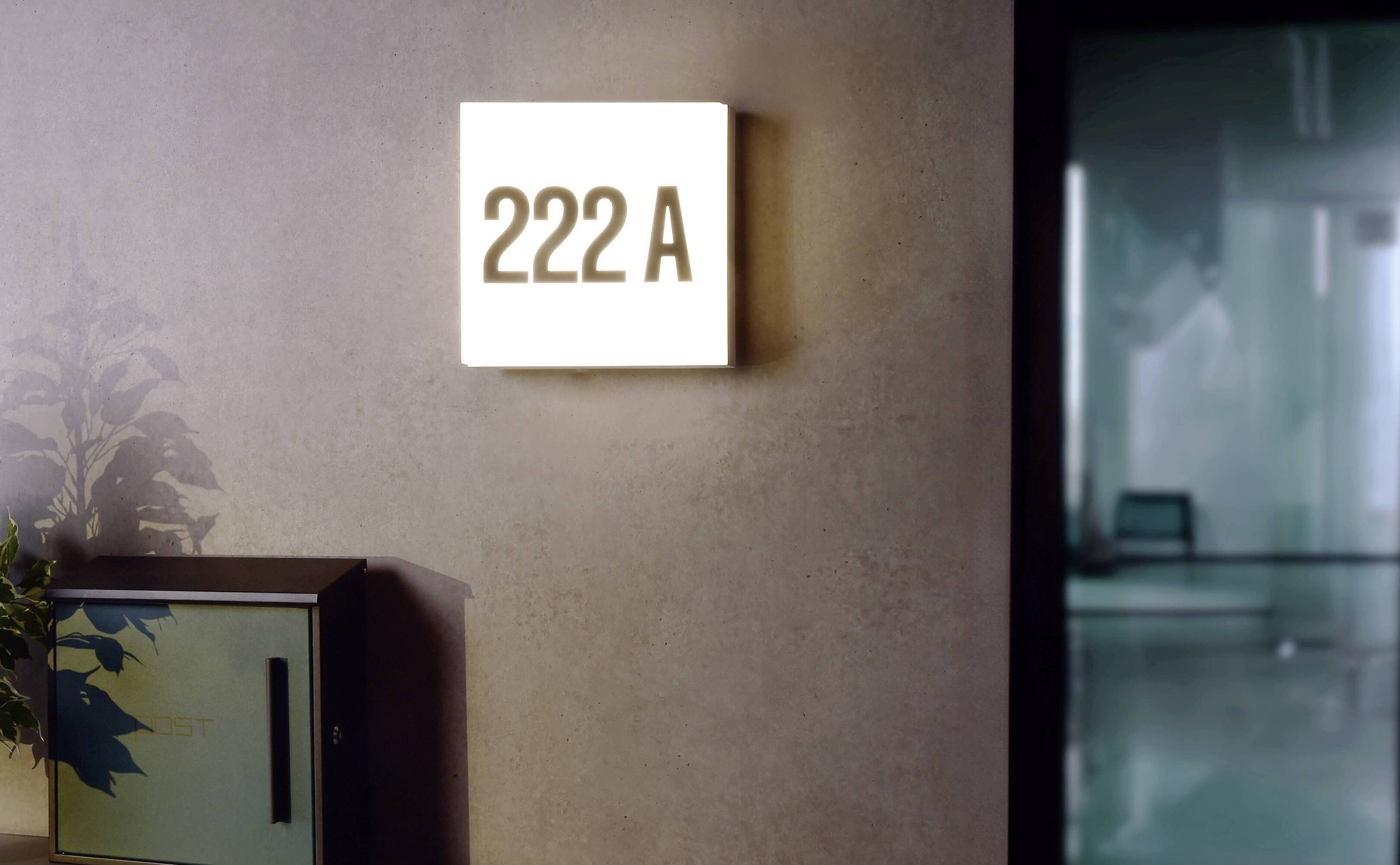 Leuchtendes Türschild mit der Aufschrift '222 A' an einer Wand, daneben eine Pflanze und ein Briefkasten.