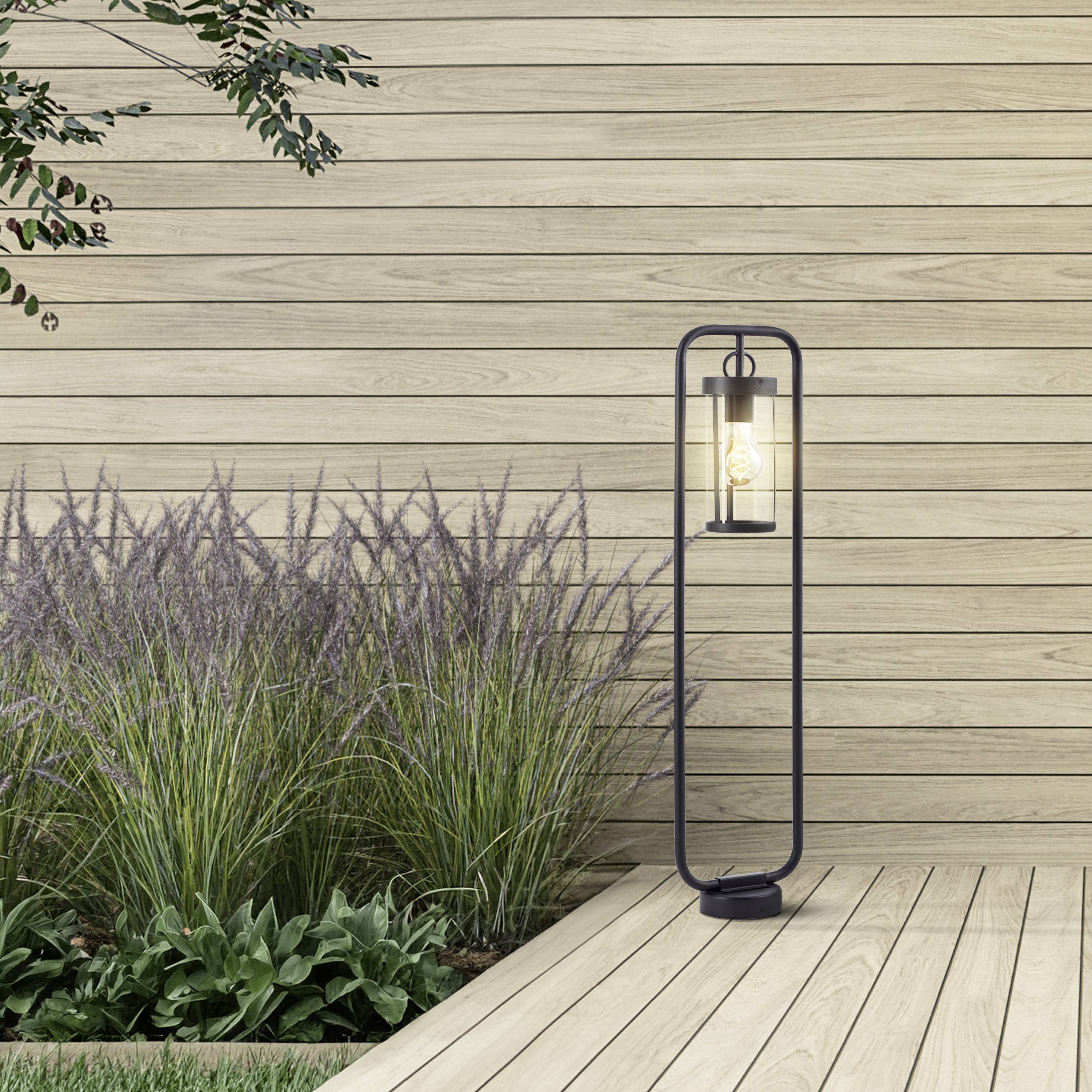 Grüne Gräser wachsen neben einer modernen Stehlampe vor einer hölzernen Wand. Eine ruhige, minimalistische Gartenszene.