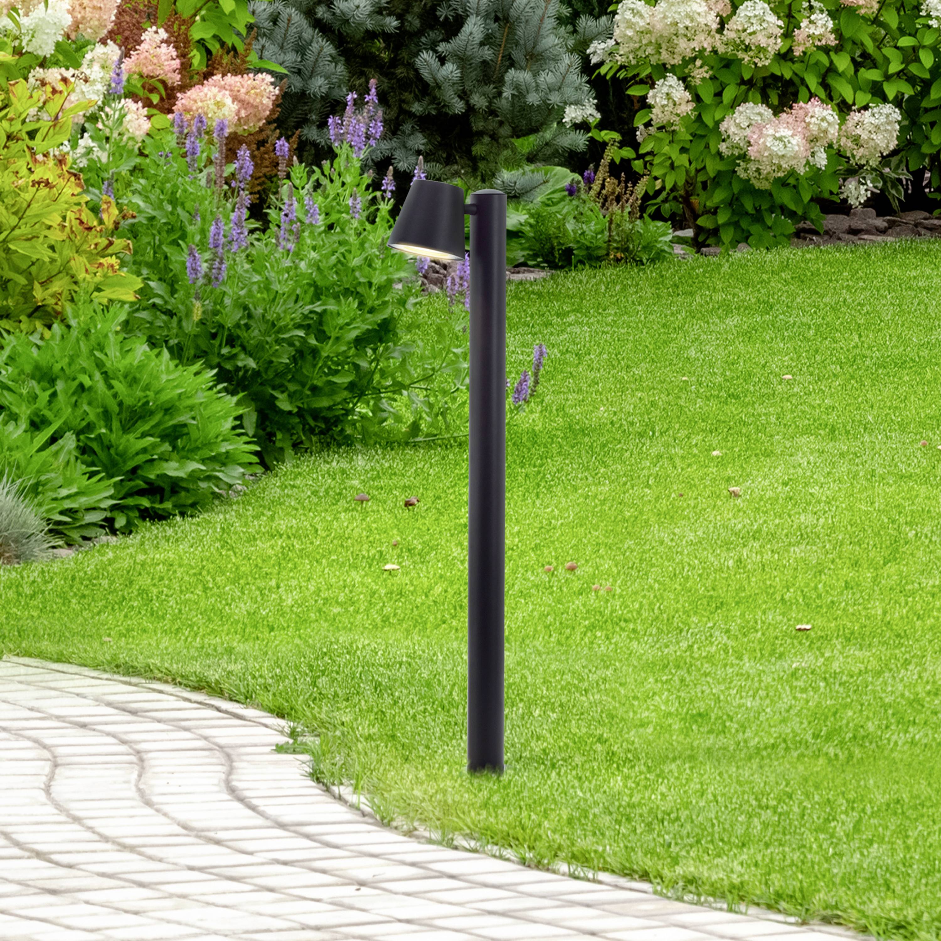 Garten mit grünem Rasen, flankiert von blühenden Pflanzen und einem geschwungenen Pflasterweg. Eine schwarze Laterne steht im Vordergrund.