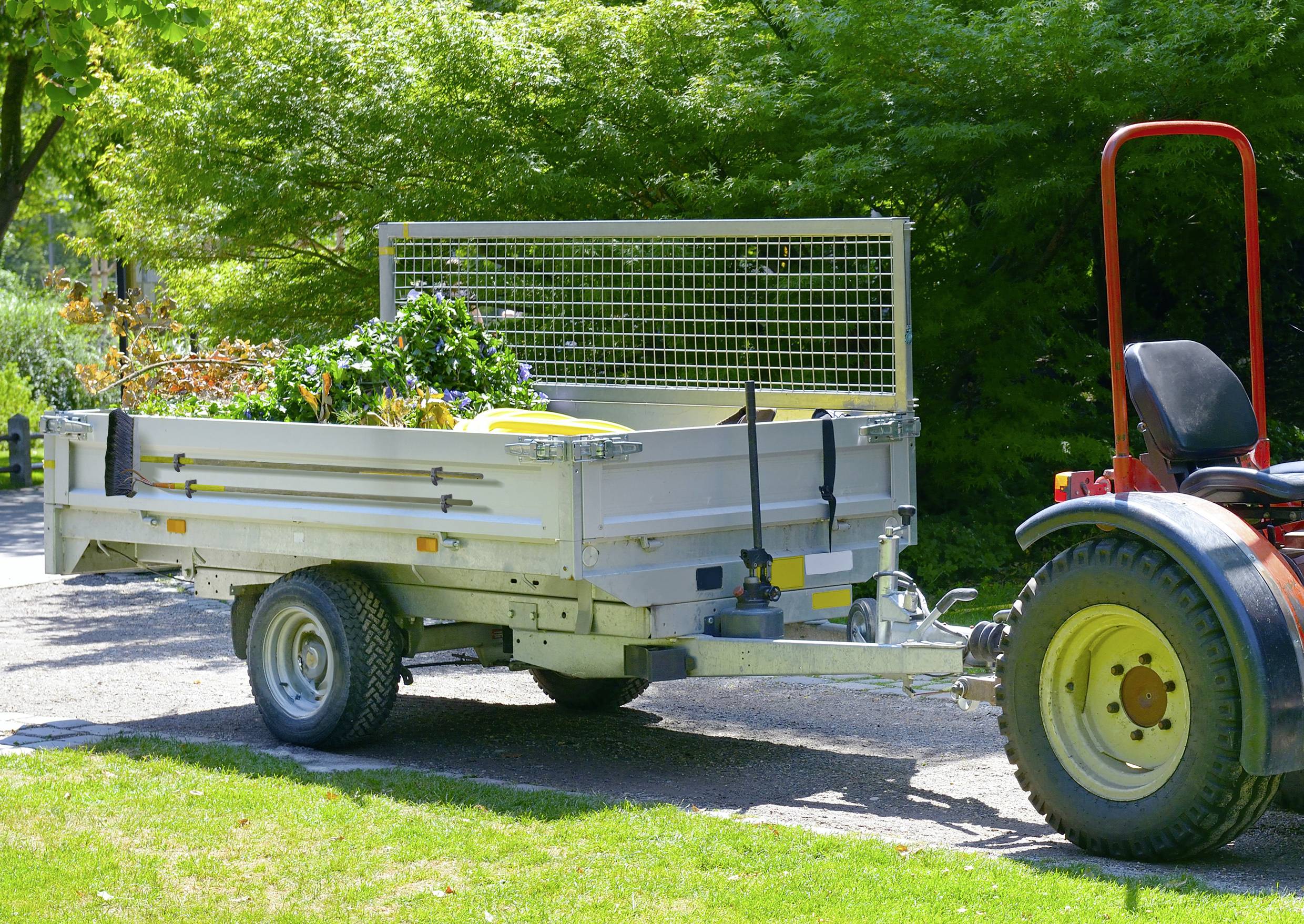 Ein kleiner Traktor zieht einen Anhänger voller Gartenabfälle auf einem sonnigen Weg, umgeben von Bäumen im Hintergrund.