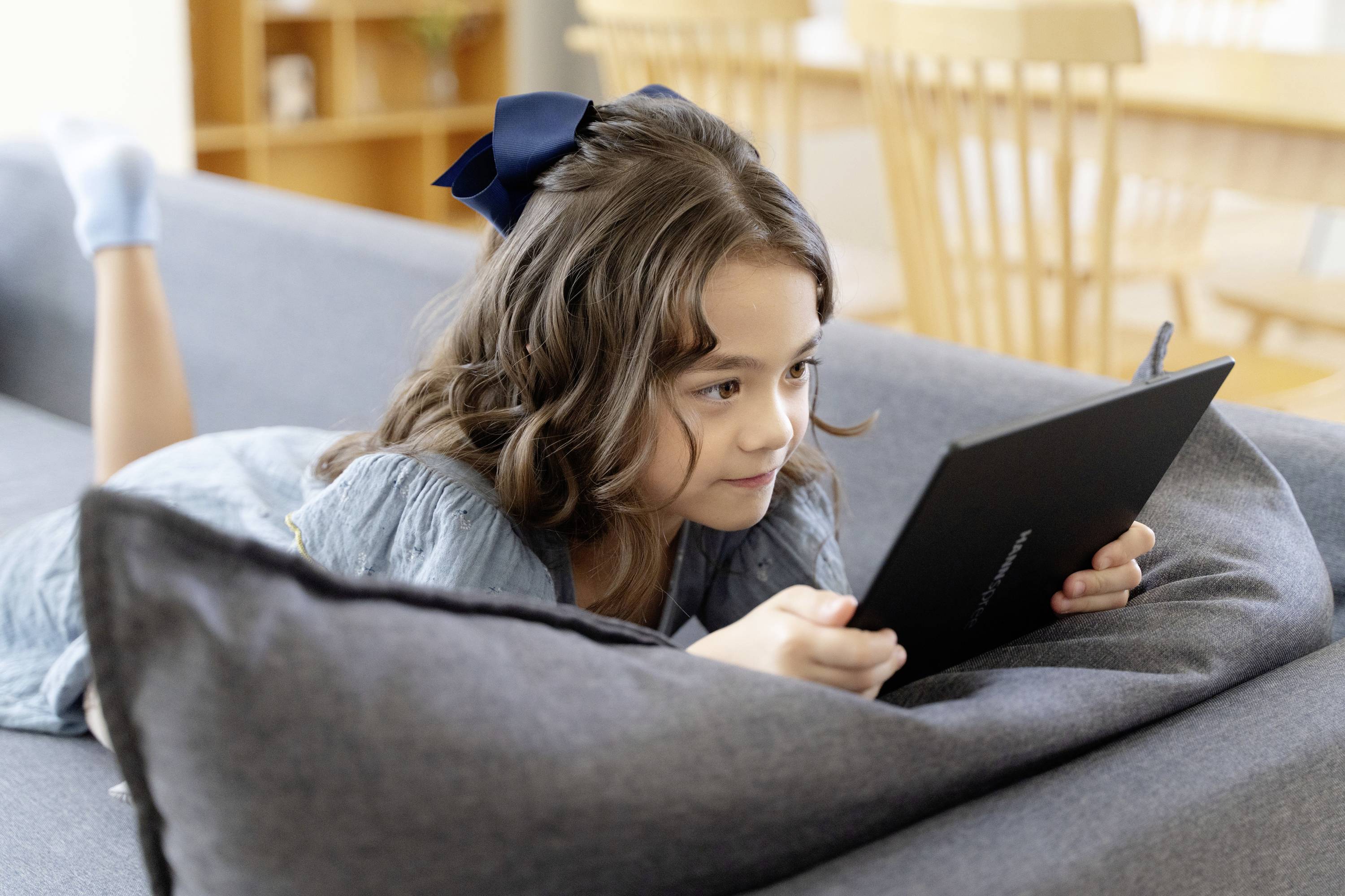 Mädchen liegt auf einem Sofa und liest oder spielt auf einem Tablet. Sie hat braunes Haar und eine Schleife als Haarband.