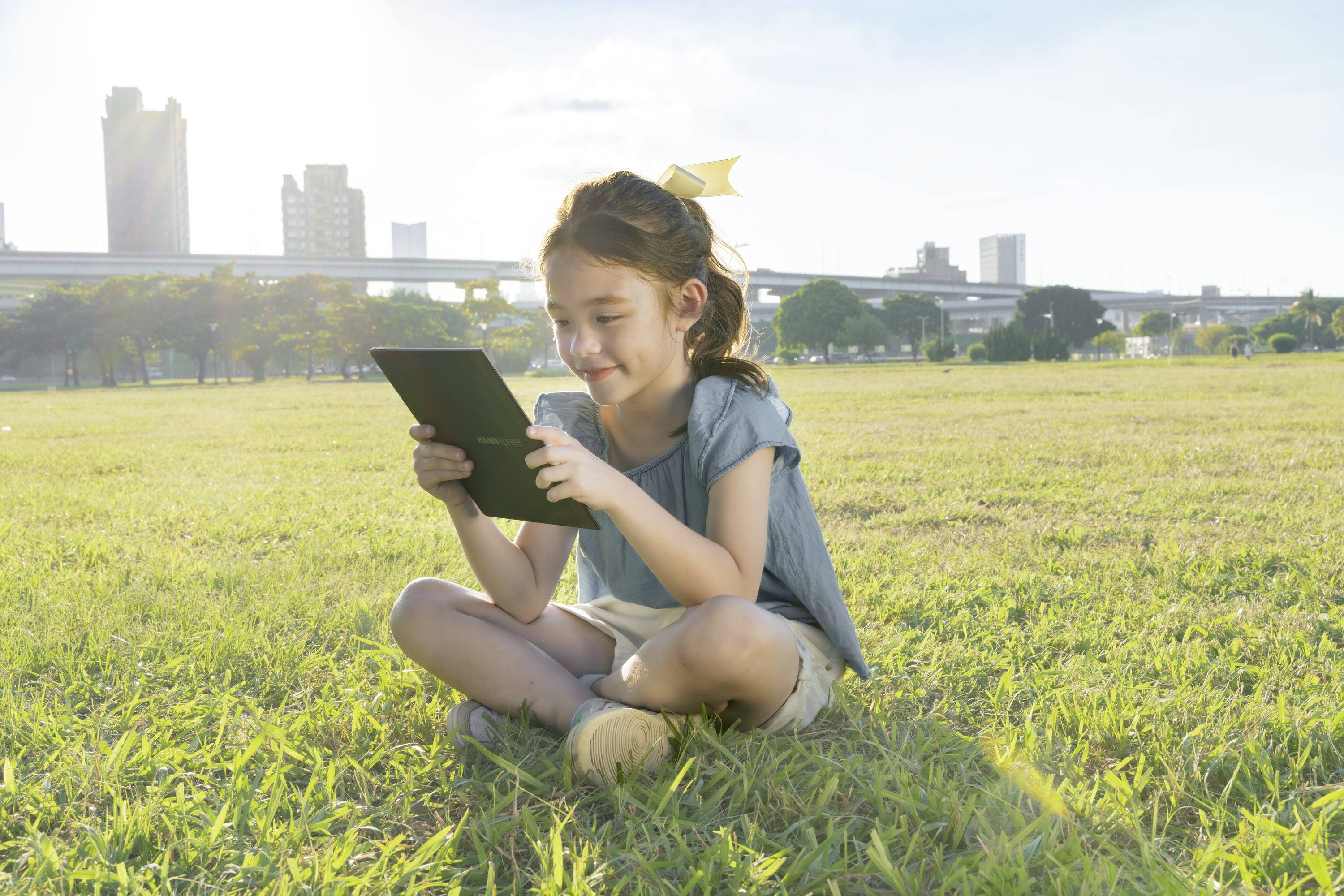 Ein Mädchen sitzt auf einer Wiese und schaut auf ein Tablet. Im Hintergrund sind Bürogebäude und ein sonniger Himmel zu sehen.