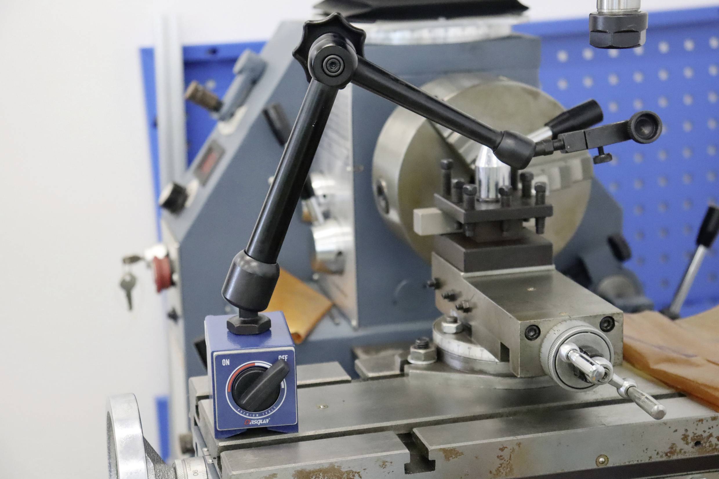 Eine Drehmaschine mit Montagearm und blauem Einstellknopf. Der Hintergrund zeigt eine blaue Werkzeugwand mit Lochmuster.