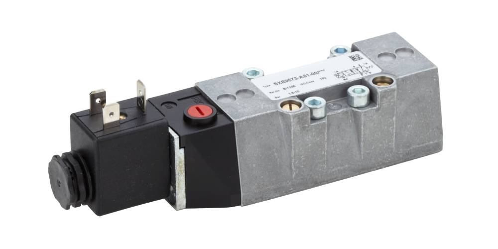 Ein Magnetventil mit silbernem, eckigem Gehäuse und schwarzem Anschlussstecker. Ein roter Schalter ist auf der Oberseite sichtbar.