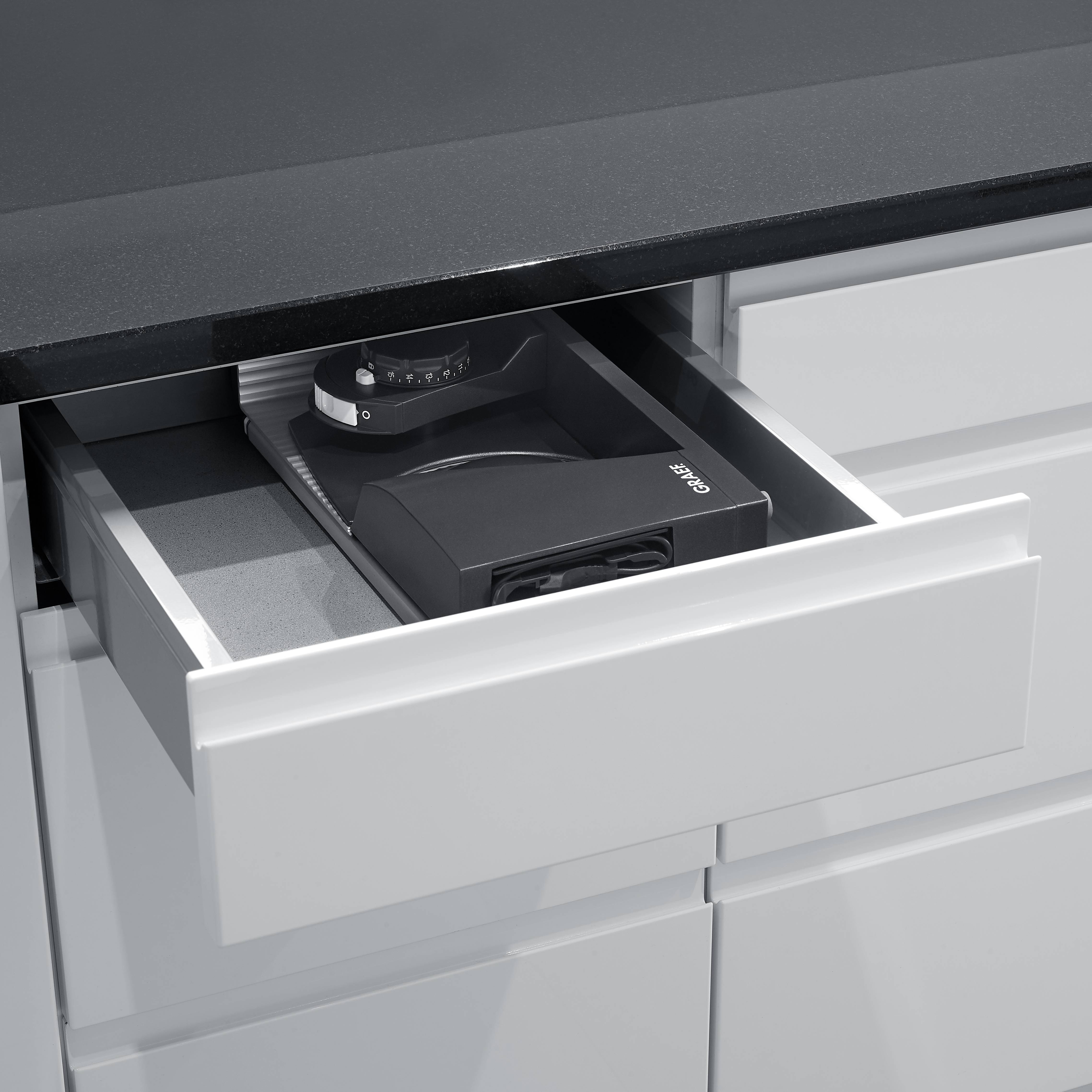 Geschlossene weiße Schublade unter einer dunklen Arbeitsplatte wird geöffnet, darin ein schwarzes Quadratgerät mit dem Wort 'QUIR'.