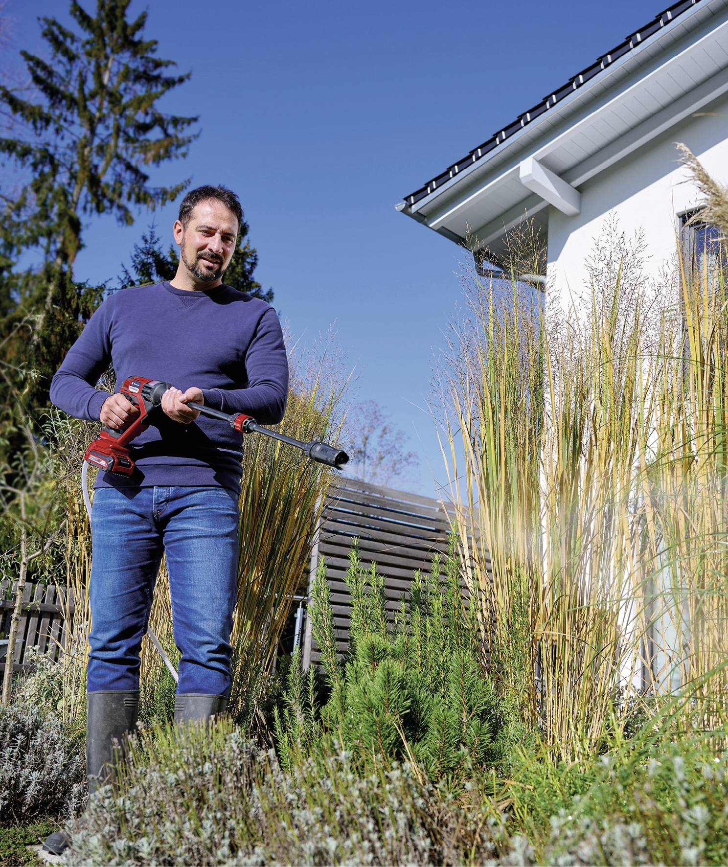 Ein Mann in Jeans und Pullover schneidet Pflanzen im Garten mit einem elektrischen Trimmer. Im Hintergrund ist ein Haus zu sehen.