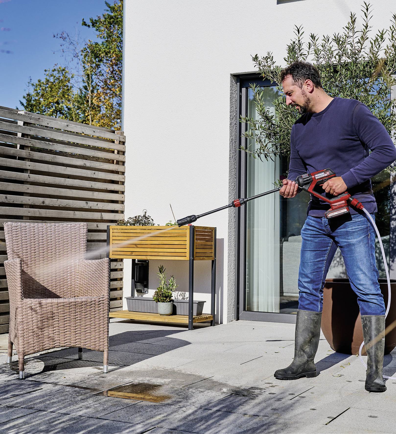 Ein Mann reinigt mit einem Hochdruckreiniger einen geflochtenen Stuhl auf einer Terrasse. Im Hintergrund sind Pflanzen und Möbel.