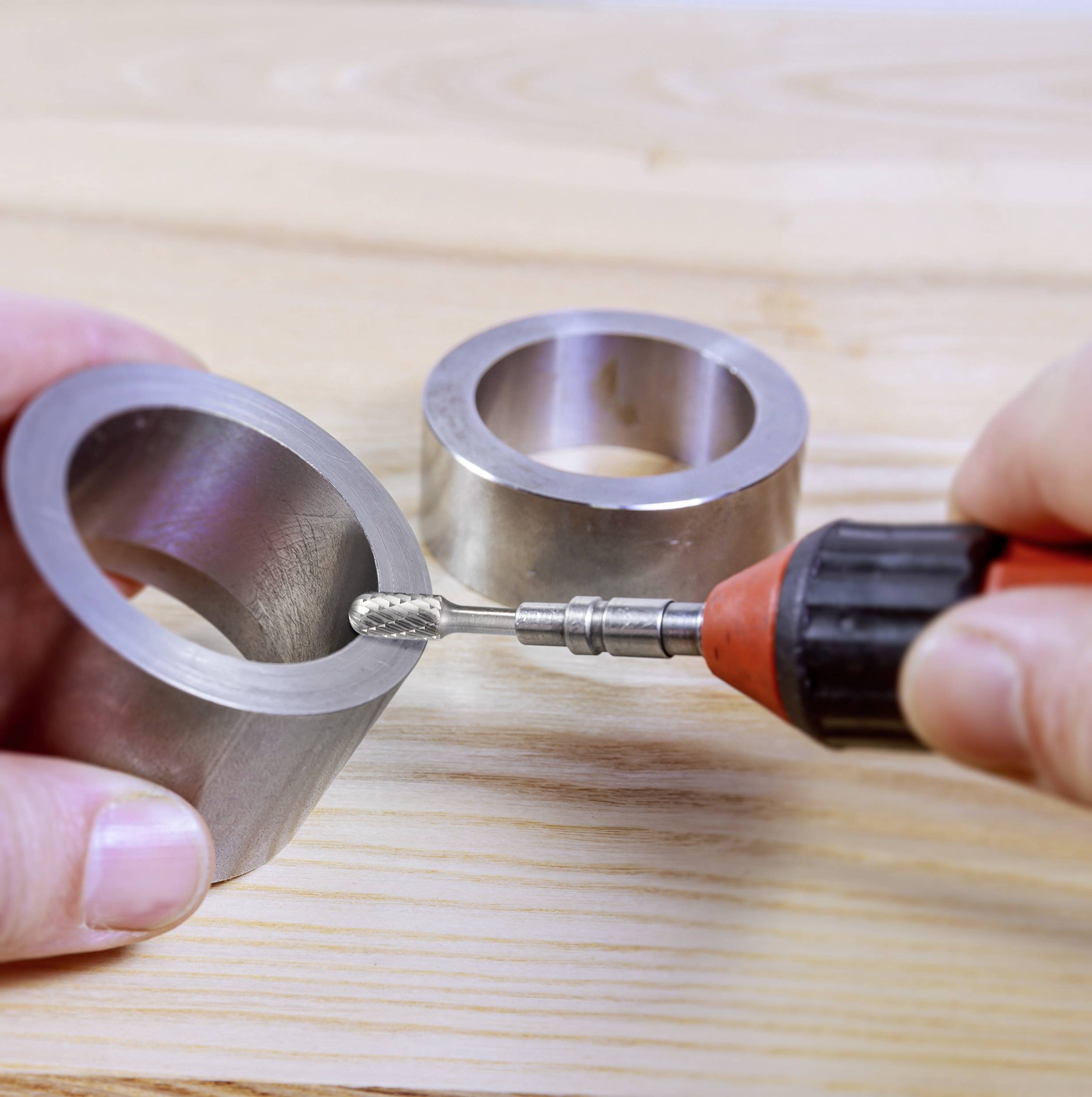 Hand hält Werkzeug in der Nähe von zwei Metallringen auf Holzoberfläche. Werkzeug wird zum Schleifen oder Bearbeiten der Ringe benutzt.
