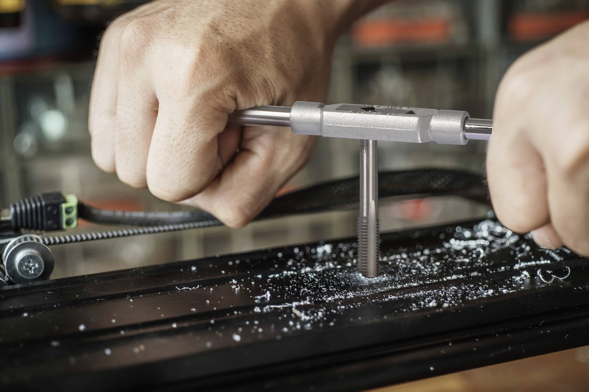 Zwei Hände schneiden mit einem Gewindebohrer ein neues Gewinde in ein Metallstück. Metallspäne sind zu sehen.