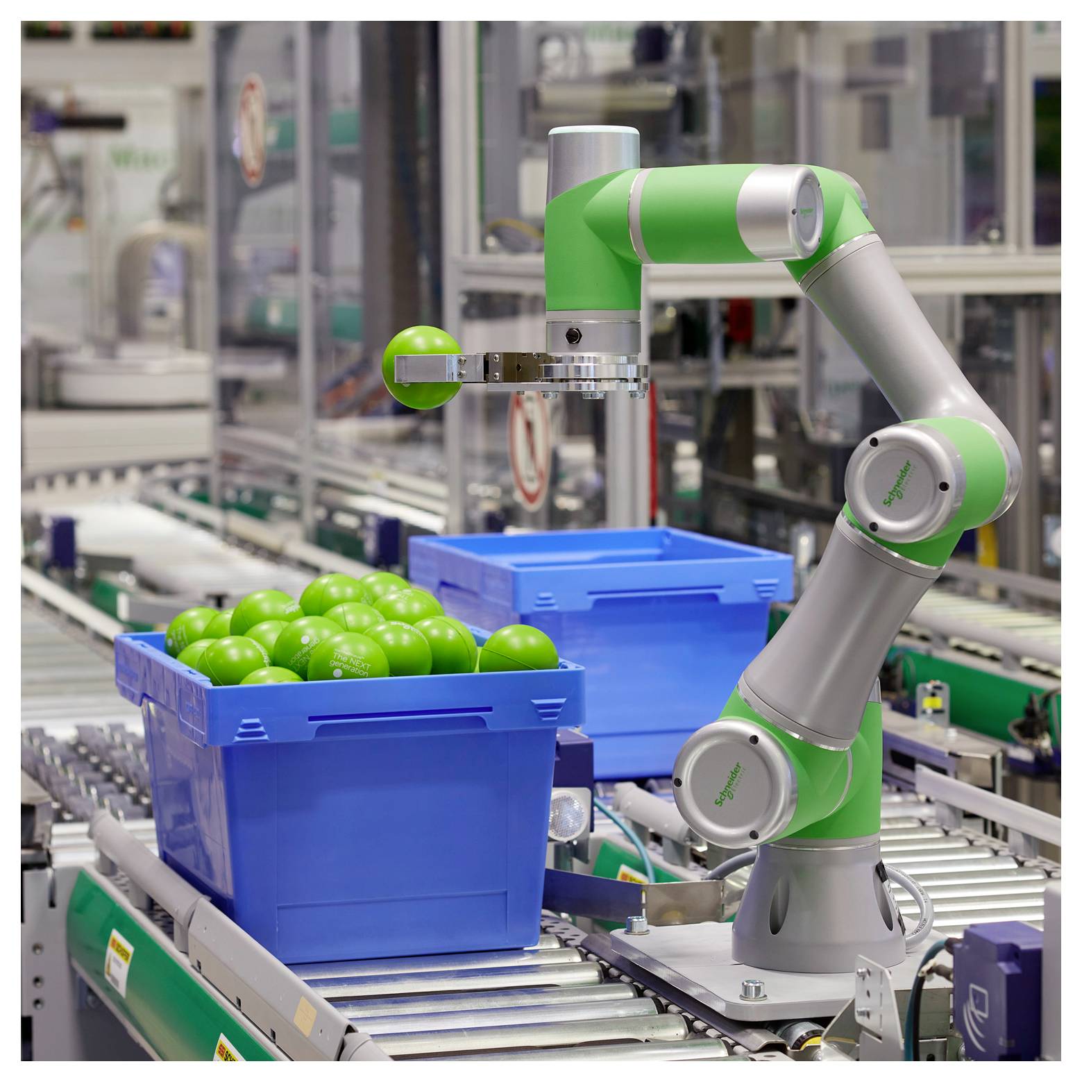 Roboterarm an einer Fließlinie, der einen grünen Ball aus einer blauen Kiste mit ähnlichen grünen Bällen aufgreift, mit Förderbandsmaschinen in einer Fabrik.