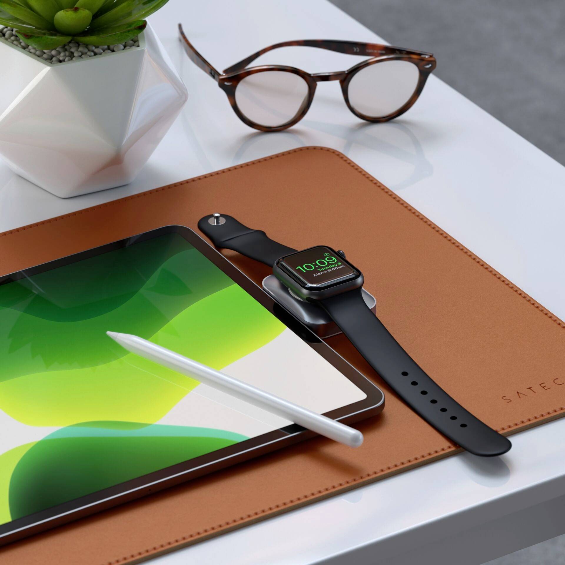 Tablet mit Stift auf Ledermatte, daneben Smartwatch mit grünem Zifferblatt; im Hintergrund Brille und Pflanze in geometrischem Topf.