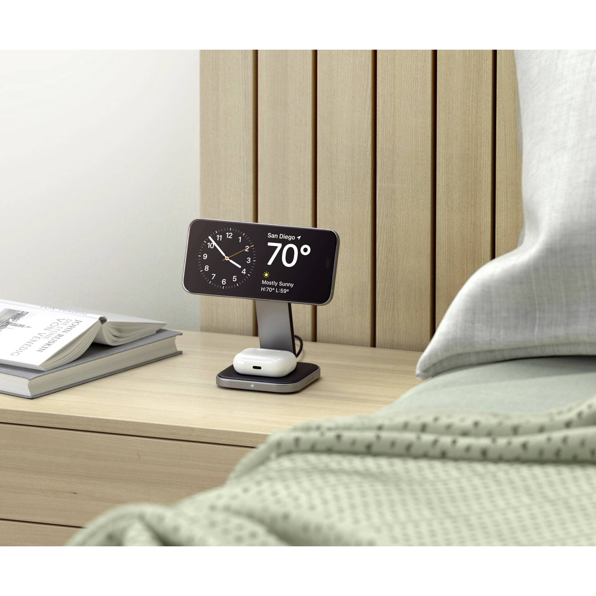 Smartphone auf Nachttisch in Ladestation, zeigt Datum, Uhrzeit und Wetter (San Diego: 70°F). Daneben ein Buch und ein ungemachtes Bett.