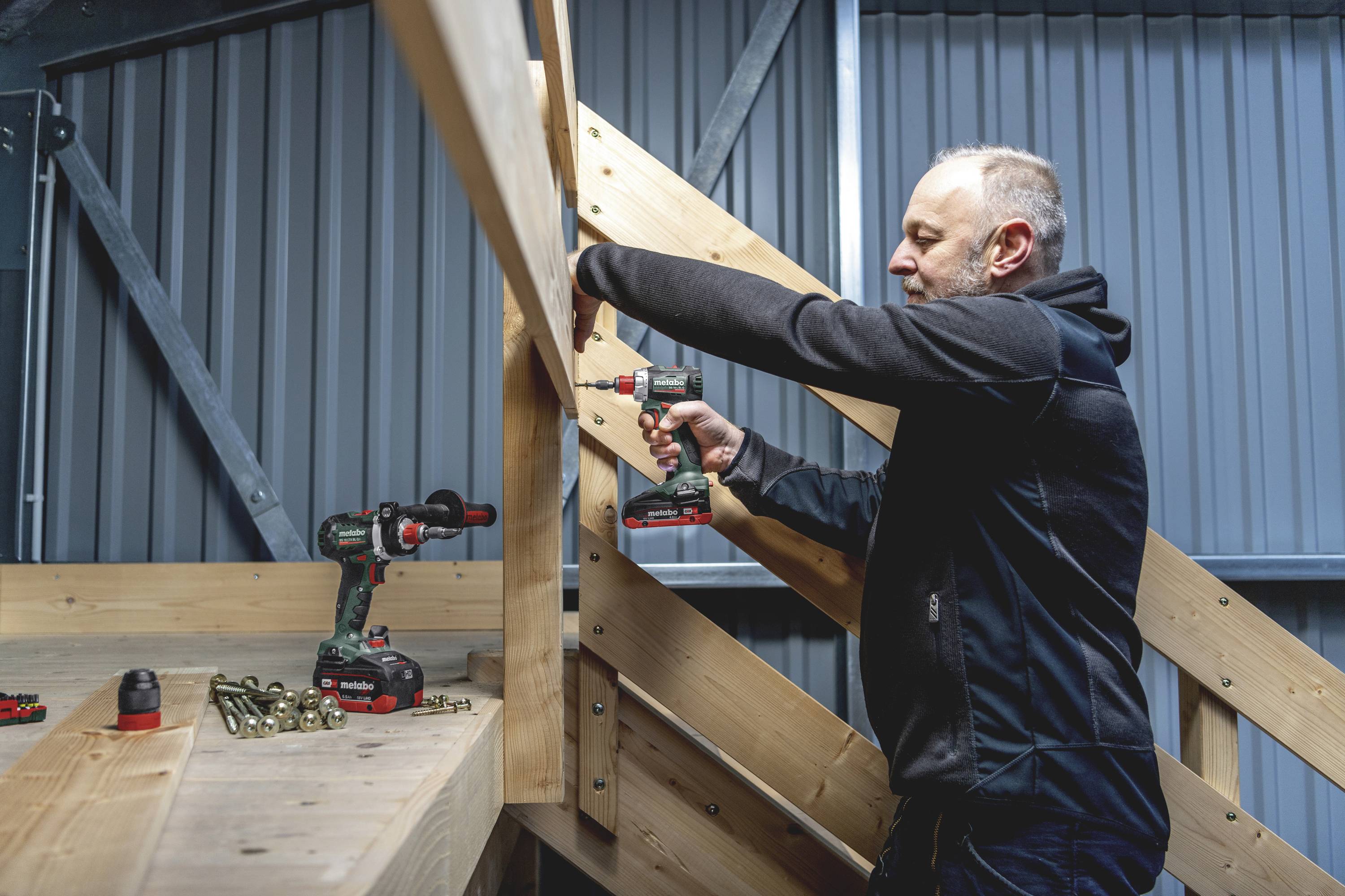 Ein Mann arbeitet mit einer Bohrmaschine an einer Holzstruktur. Auf dem Tisch liegen weitere Werkzeuge und Schrauben bereit.