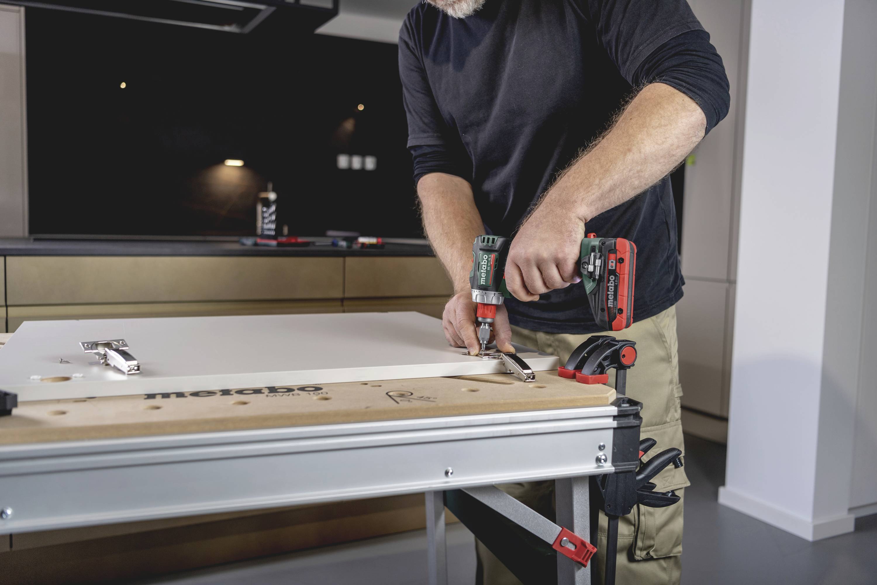 Eine Person benutzt eine Bohrmaschine, um eine Schraube in ein Holzbrett auf einem Tisch in einer modernen Küche zu befestigen.