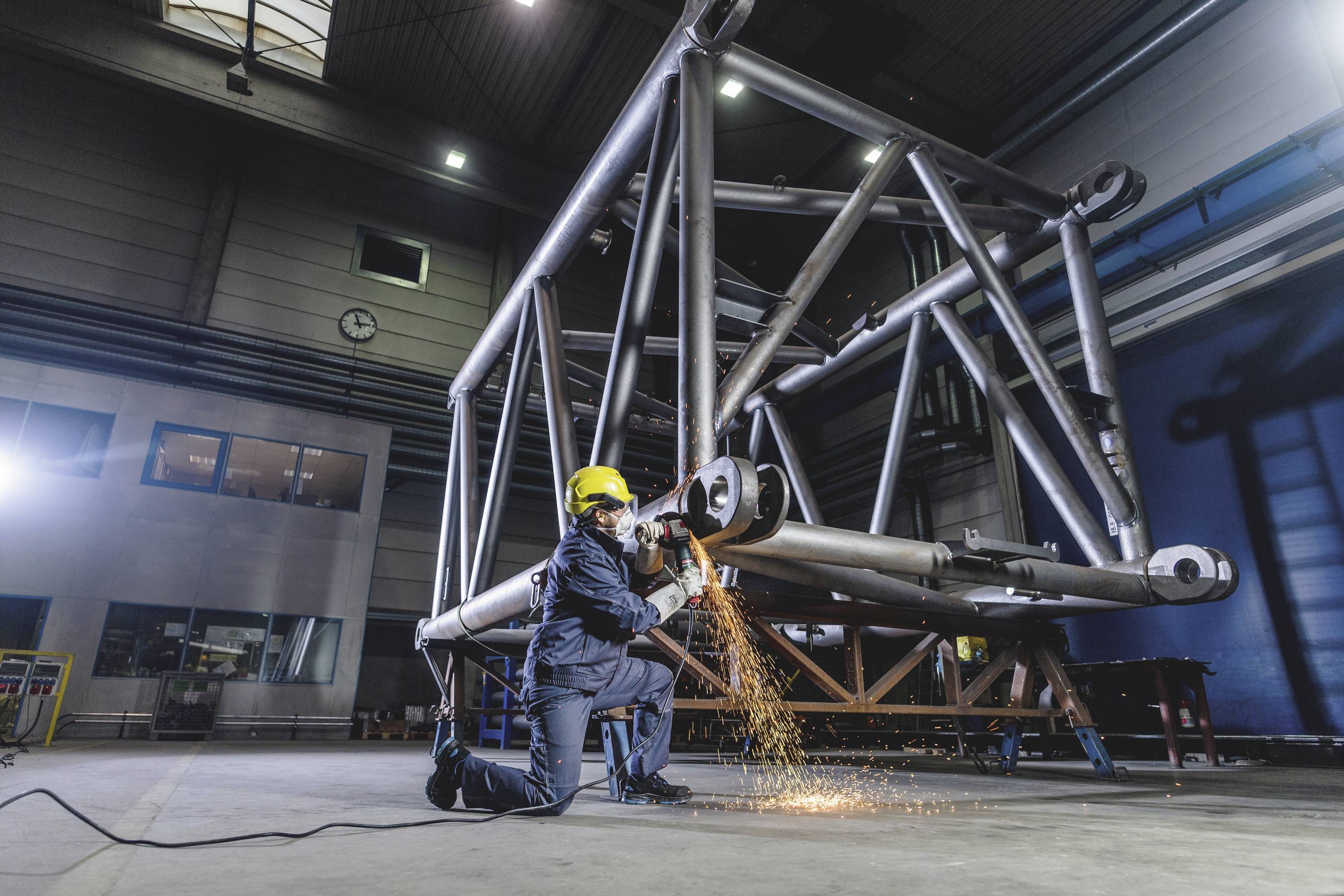Ein Arbeiter mit Schutzhelm schweißt an einer großen Metallstruktur in einer Werkshalle. Funken fliegen, Umgebung industriell.