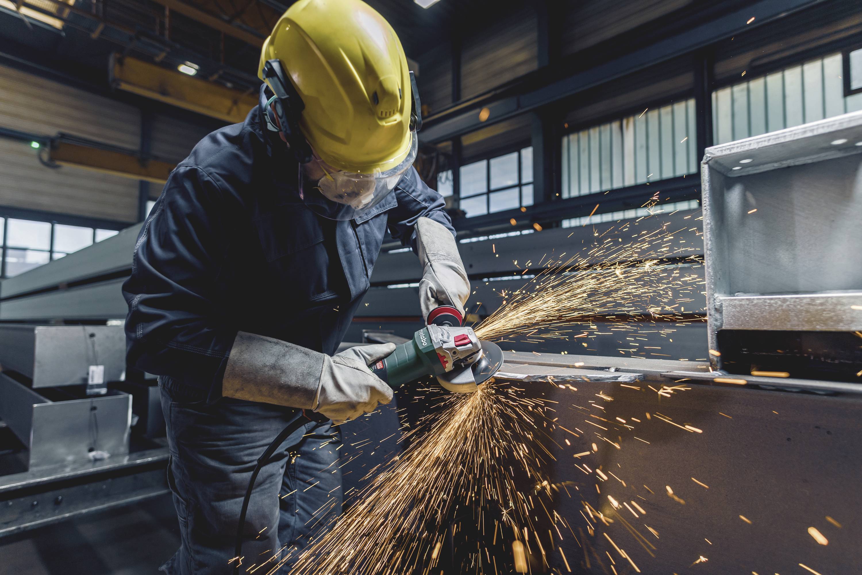 Person mit Schutzhelm und Brille schleift Metall mit einem Winkelschleifer in einer Fabrik. Funken fliegen, Sicherheitsausrüstung getragen.