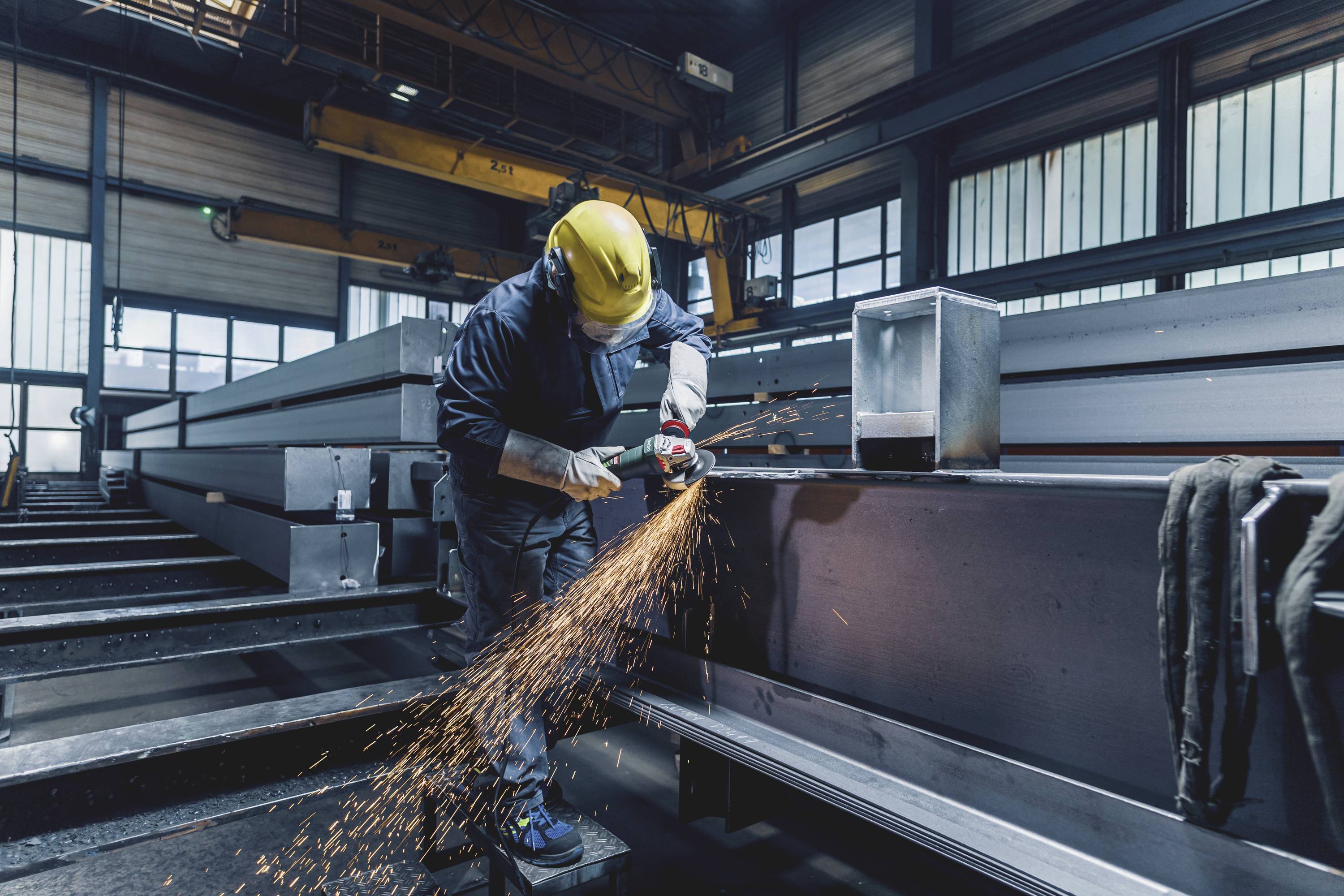 Ein Arbeiter in Schutzkleidung schleift Metallträger in einer Fabrikhalle. Funken fliegen während des Schleifens.