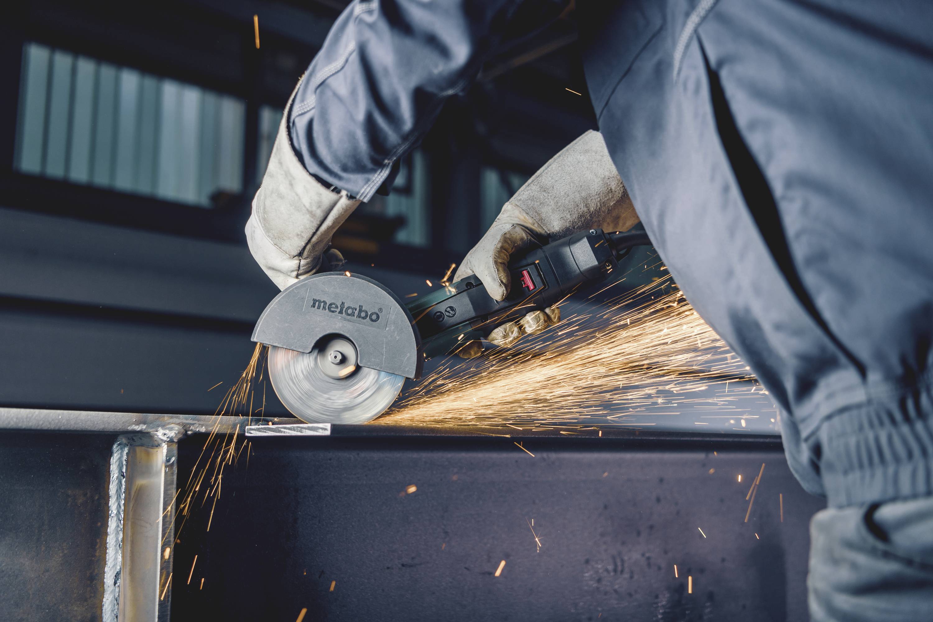 Eine Person schneidet mit einer Kreissäge Metall, Funken fliegen. Sie trägt Schutzkleidung, darunter Handschuhe.