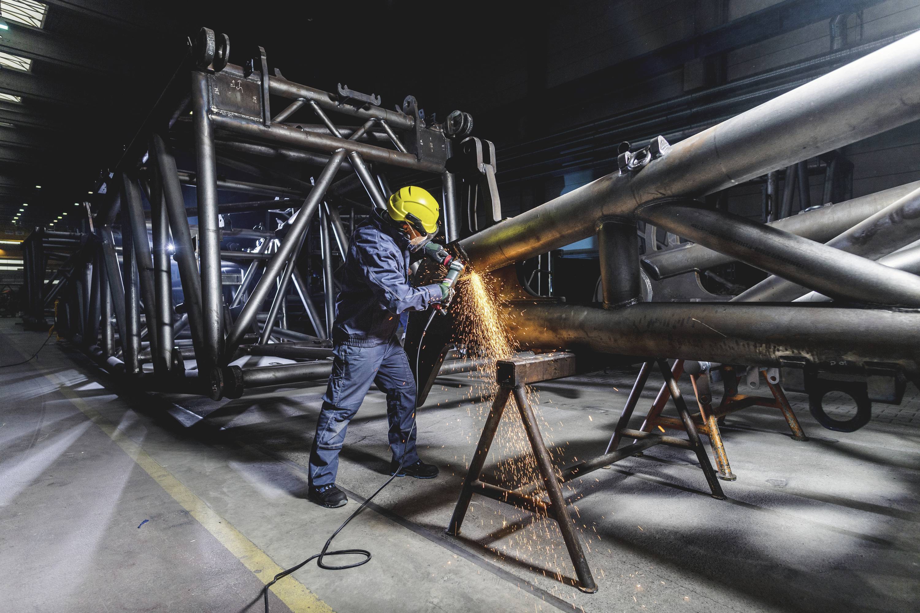 Ein Arbeiter mit Schutzhelm schleift Metallrohre in einer Fabrikhalle. Funken fliegen vom Schleifgerät ab.