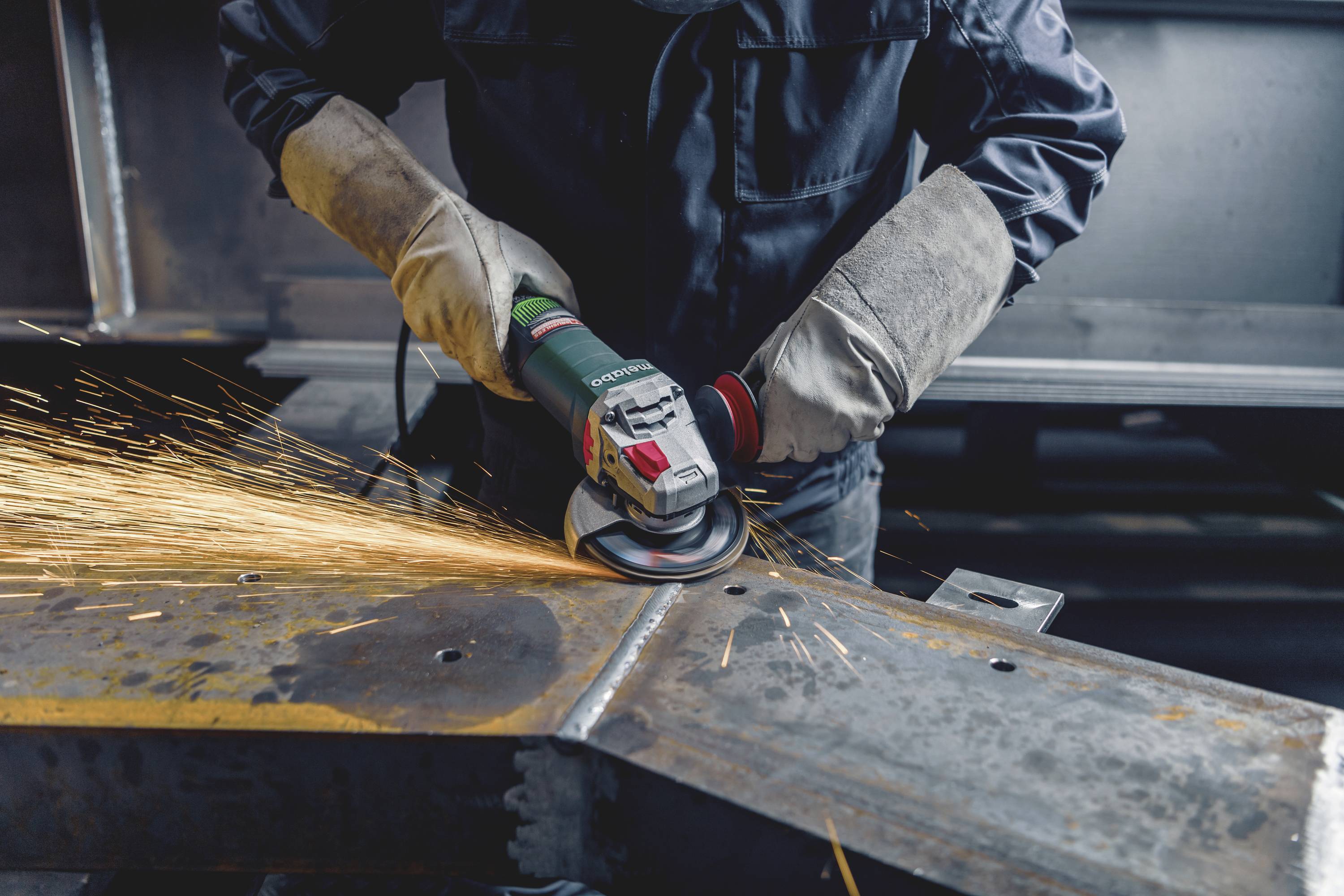 Arbeiter schleift metallisches Werkstück mit Winkelschleifer, Funken sprühen. Trägt Schutzkleidung und Arbeitshandschuhe.