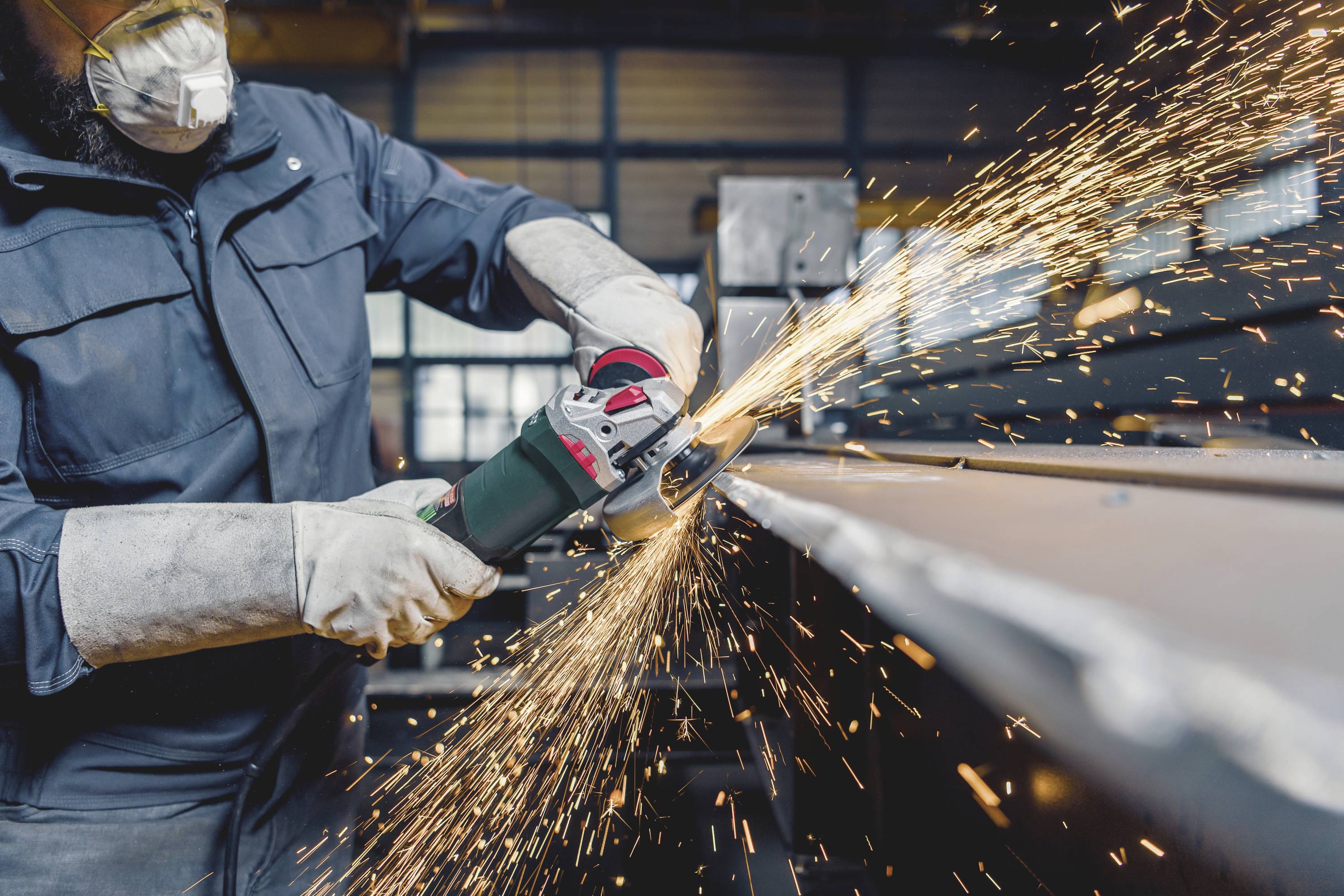 Eine Person in Schutzkleidung schleift mit einem Winkelschleifer Metall, Funken sprühen. Arbeitet in einer Werkstattumgebung.