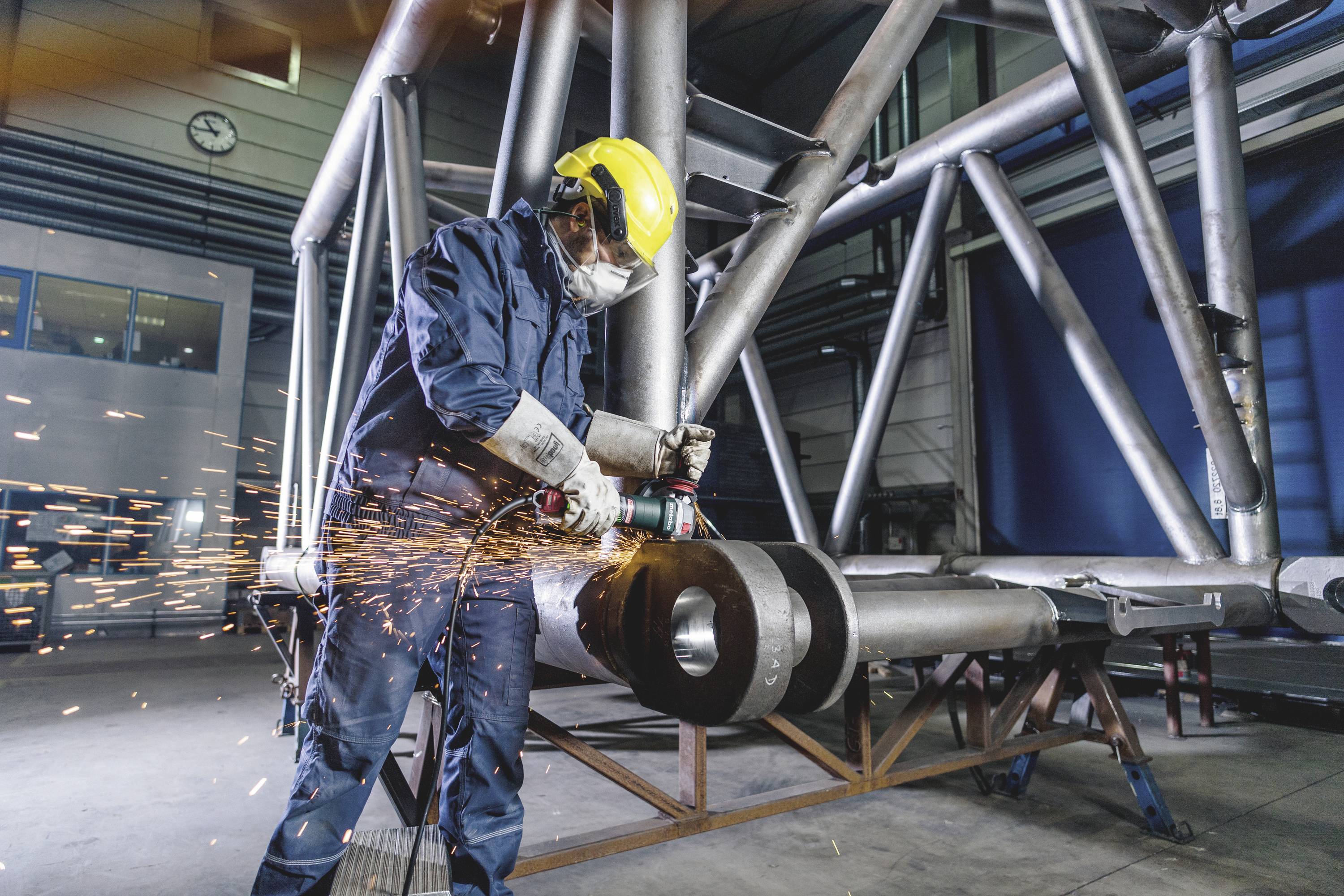 Ein Arbeiter in Schutzkleidung schleift Metall in einer Fabrikhalle, Funken fliegen herum. Im Hintergrund sind Stahlträger sichtbar.